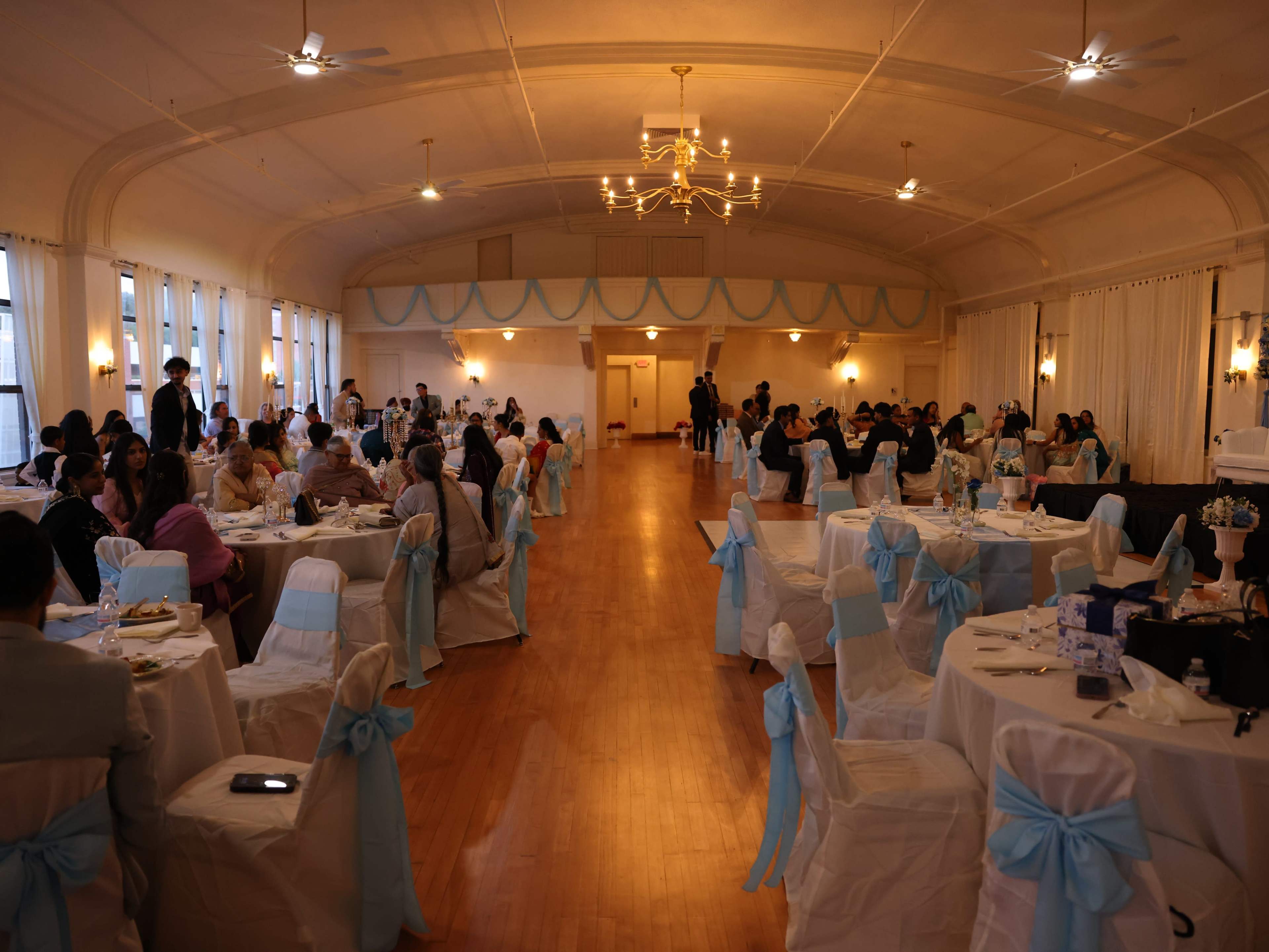 The image shows a large banquet hall with multiple tables set for dining, decorated with white and light blue chair sashes, and filled with guests engaged in conversation.