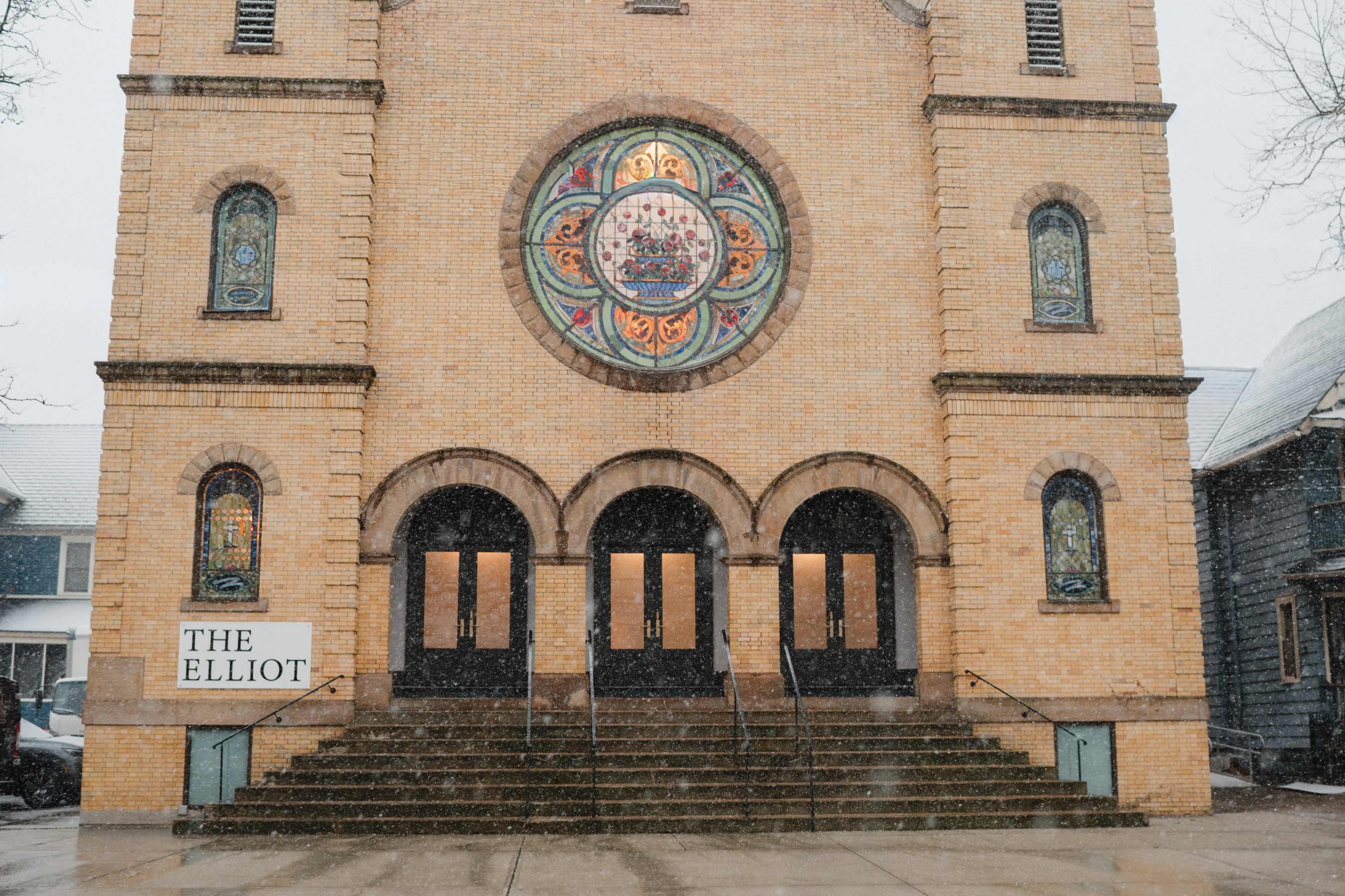 The image shows a brick building with a large stained glass window at the center, featuring stairs leading up to a grand entrance, and a sign that reads "THE ELLIOT" at the base.