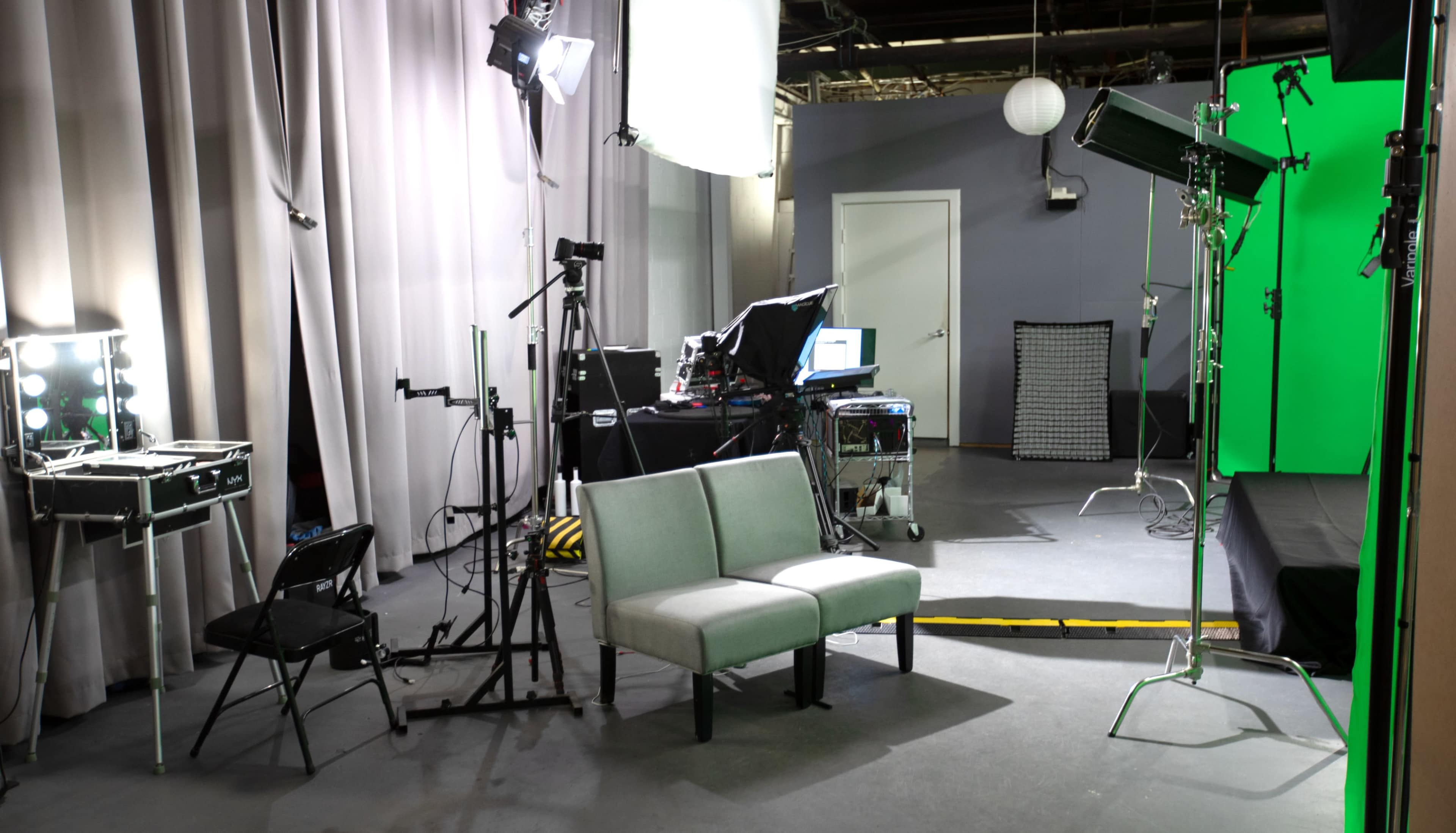 The image shows a studio setup featuring a green screen, lighting equipment, and a gray couch in the center.