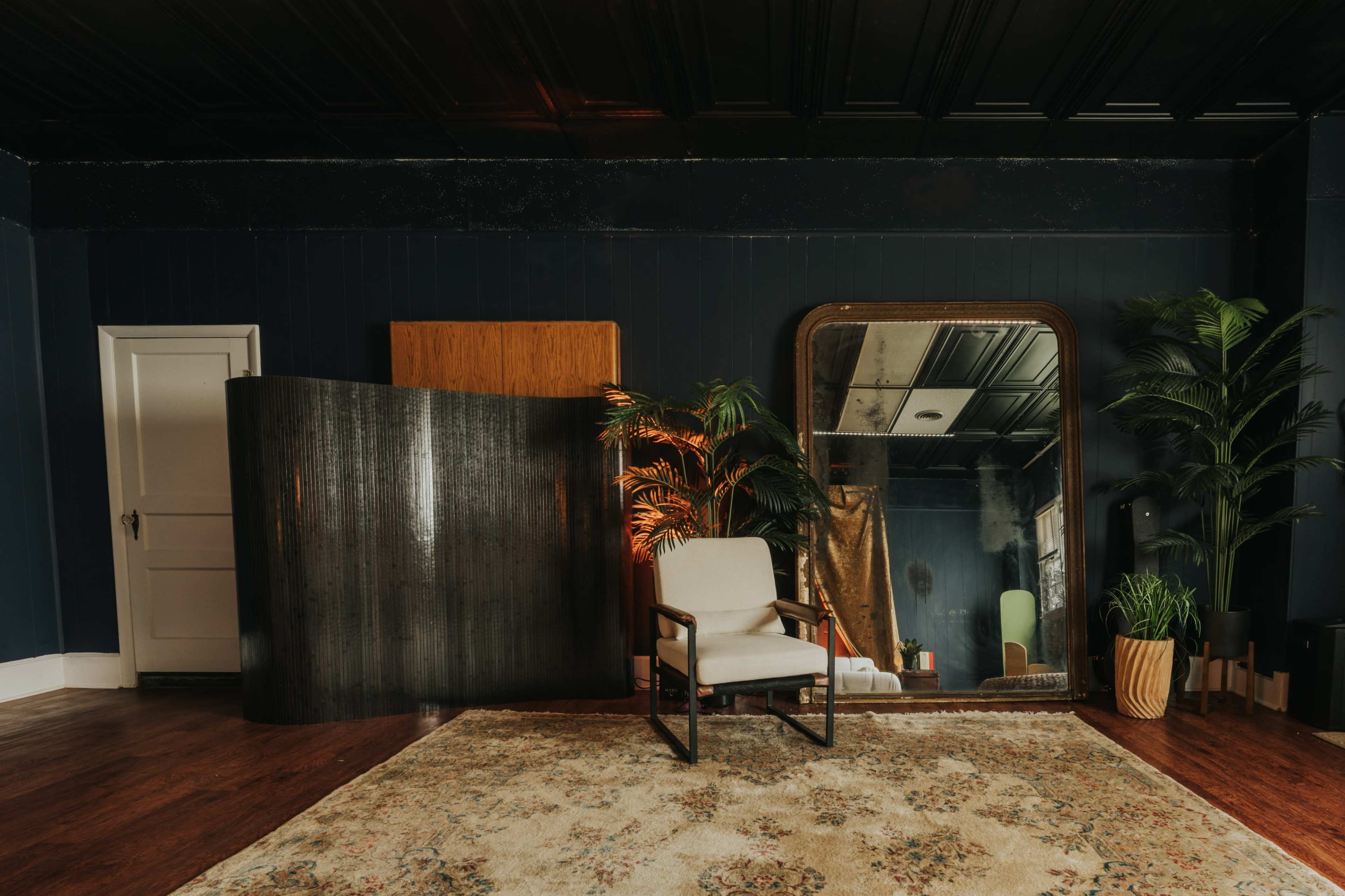 A styled interior room featuring a single chair, large mirror, decorative plants, and a patterned rug on the wooden floor.