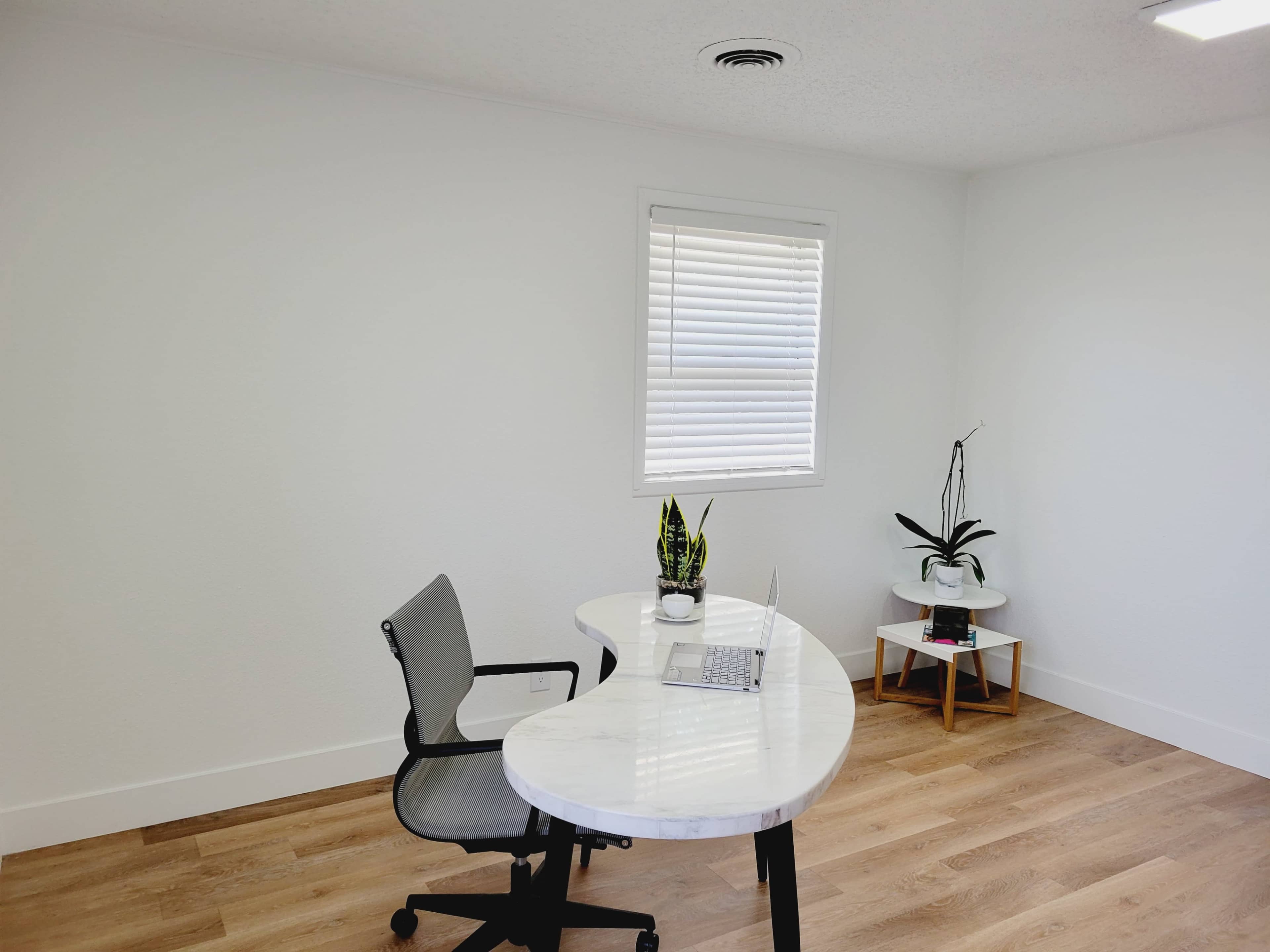 An office space features a white desk with a laptop, a chair, and potted plants next to a window.