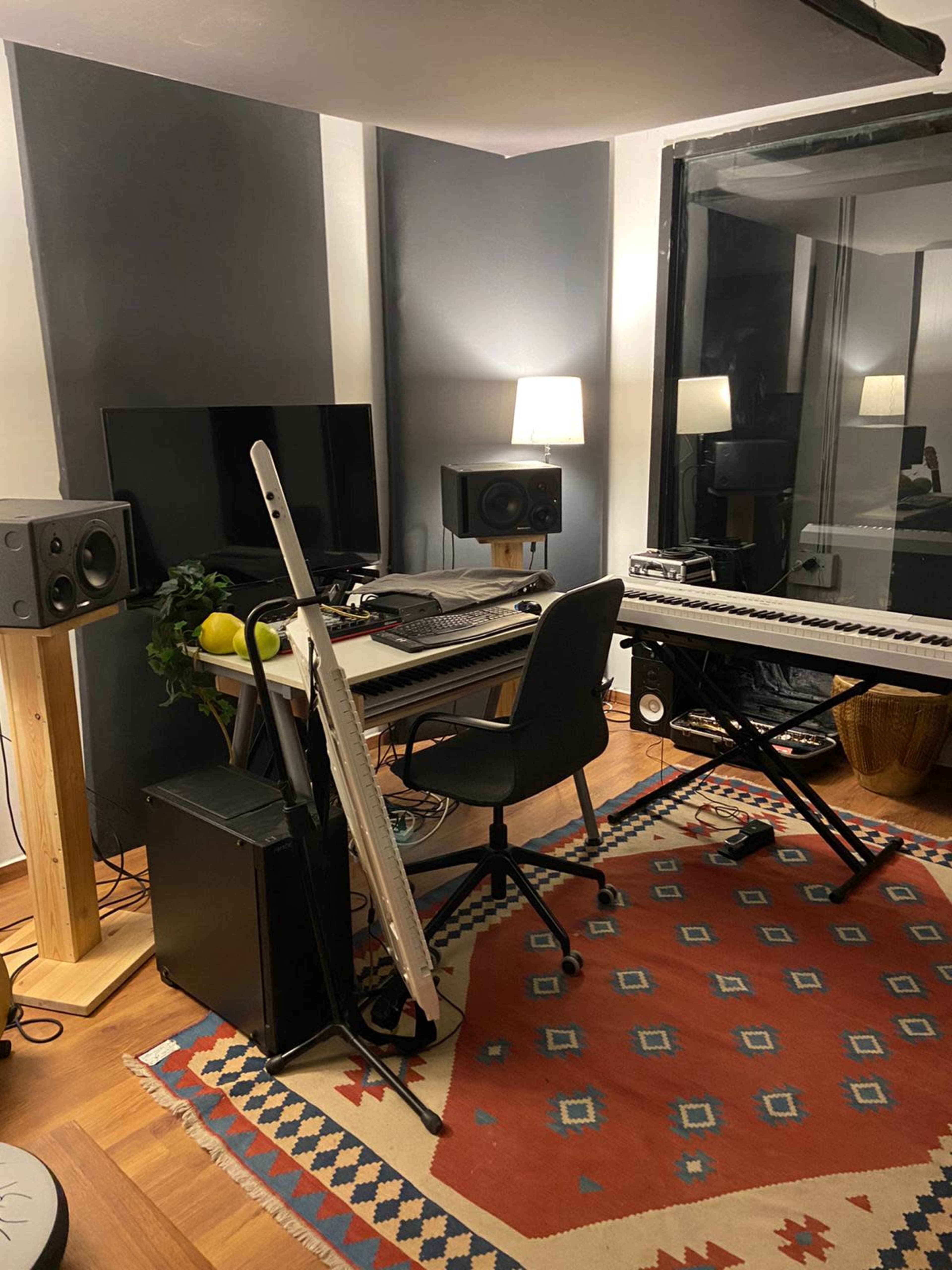 A music studio setup featuring a desk with a keyboard, speakers, a television, and a lamp, all arranged on a patterned rug.