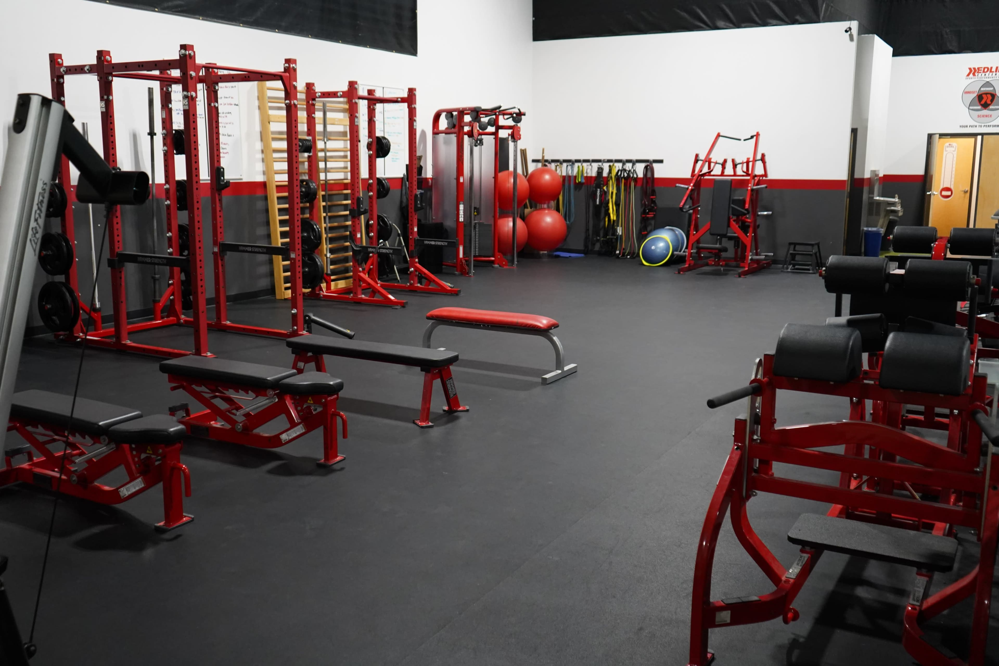 The image shows a fitness gym with various exercise equipment, including weight benches, squat racks, and exercise balls arranged in a spacious area.