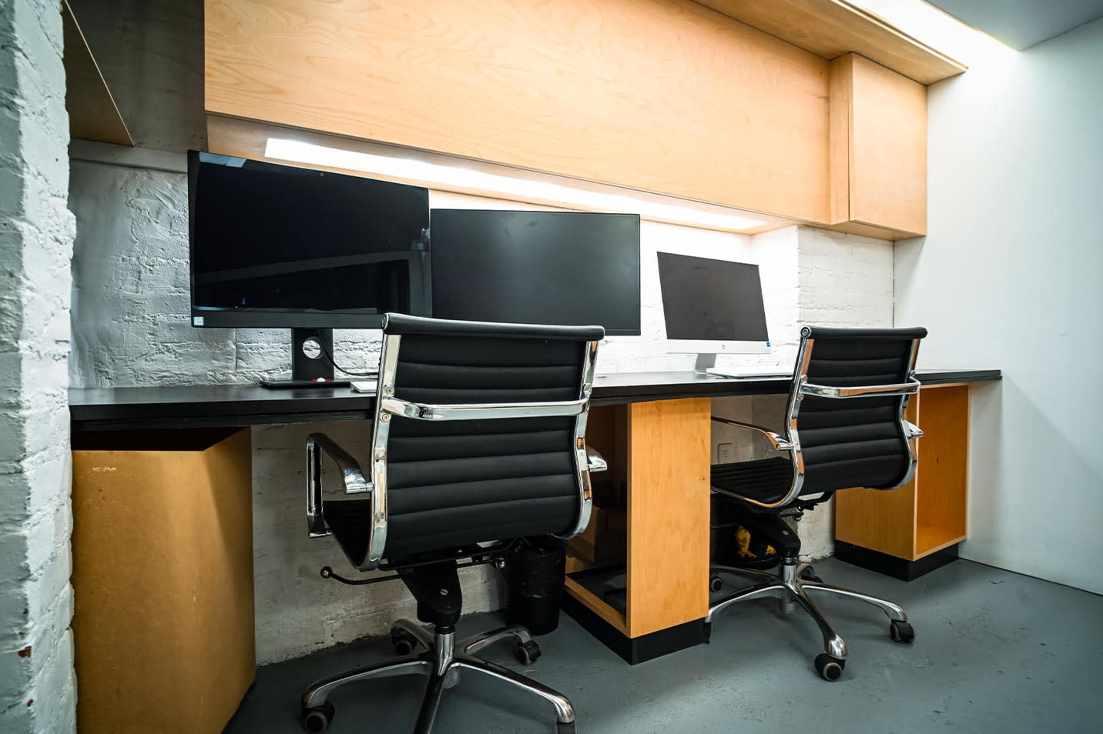 The image shows a minimalist office space with two black office chairs and two monitors on a wooden desk, located against a white brick wall.