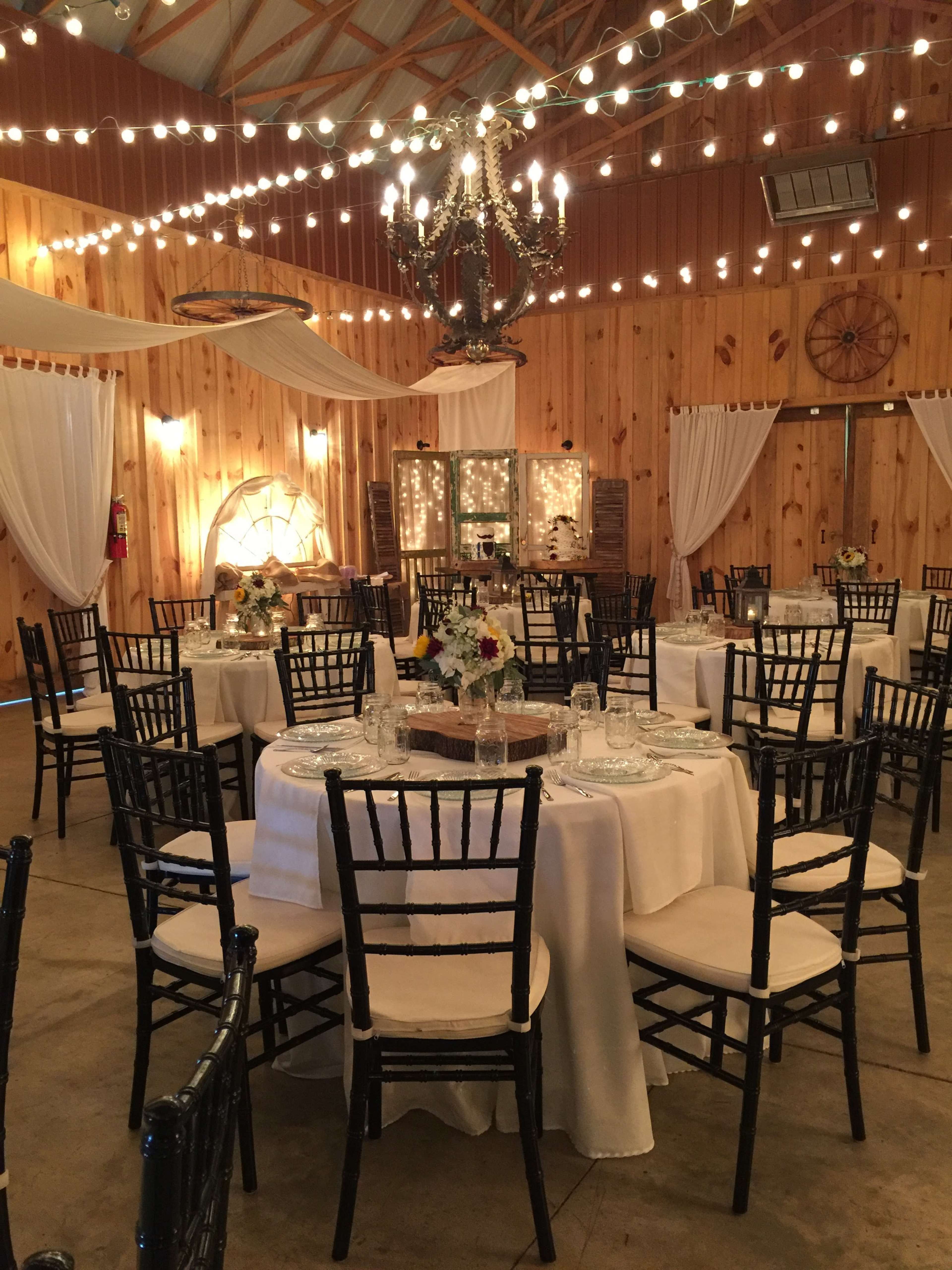 A banquet hall with round tables covered in white tablecloths, adorned with floral centerpieces, and surrounded by black Chiavari chairs under a canopy of string lights.