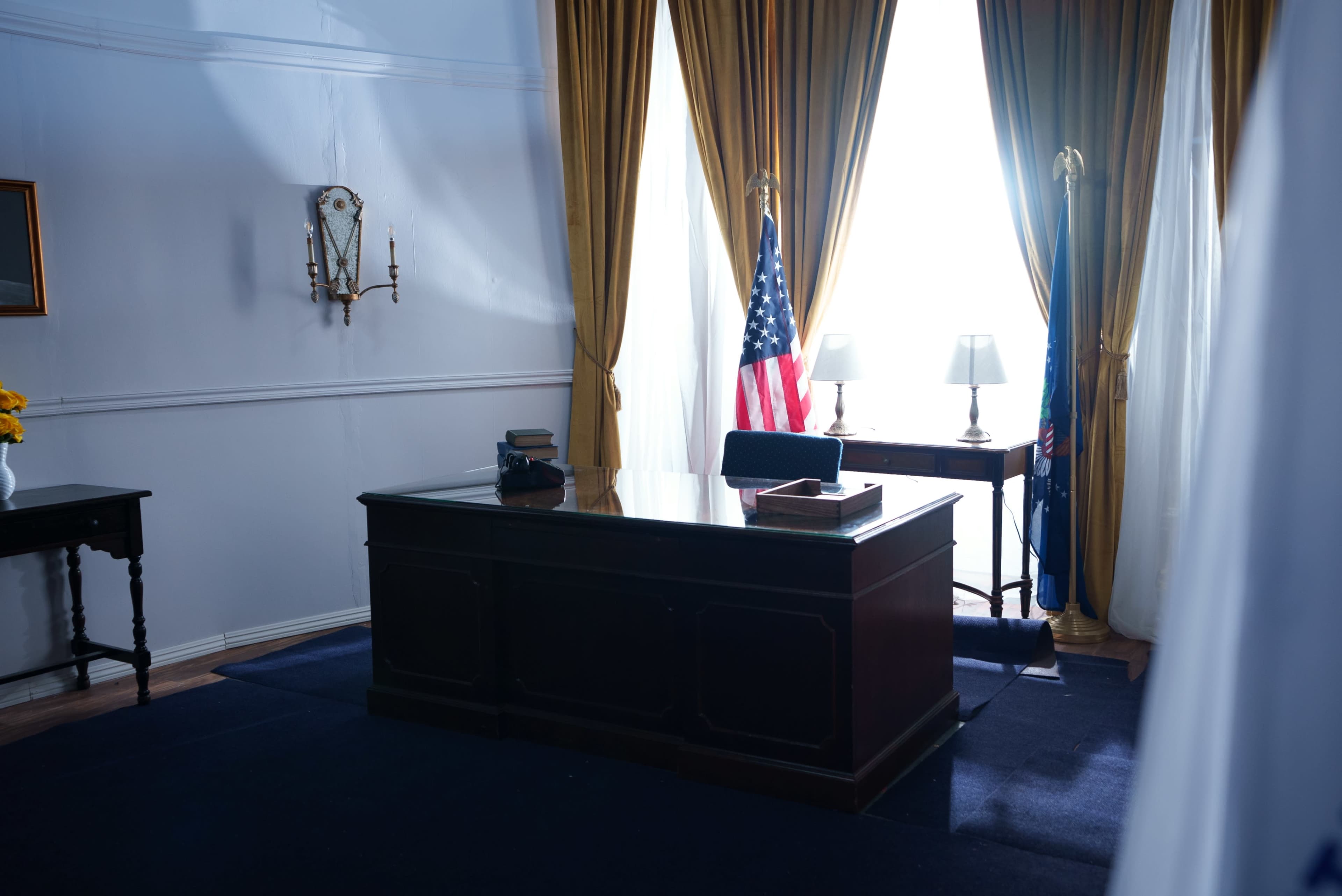 A formal office space with a wooden desk, two lamps, an American flag, and large windows draped with yellow curtains.