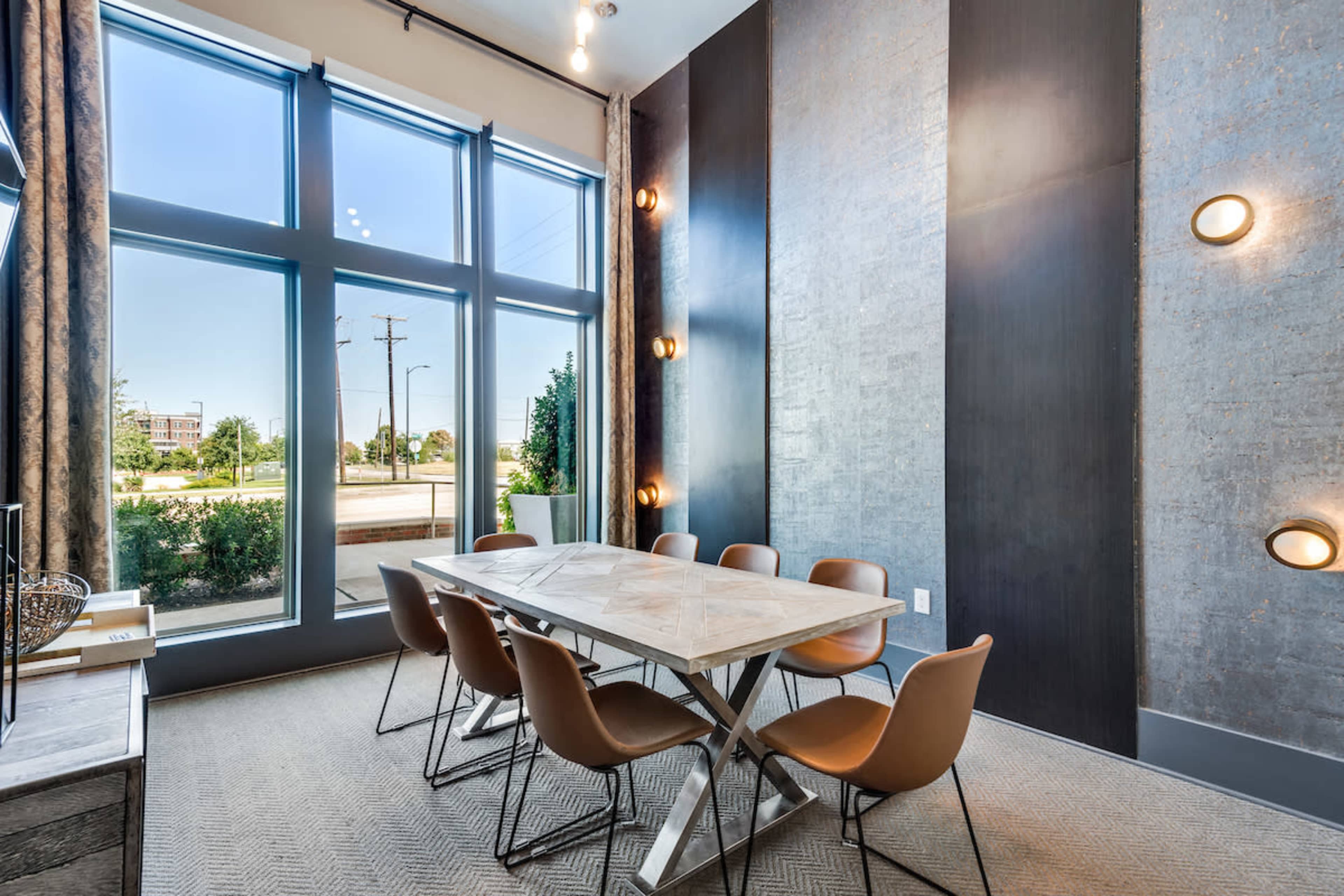 A meeting room features a large table surrounded by eight chairs, with large windows allowing natural light to enter.