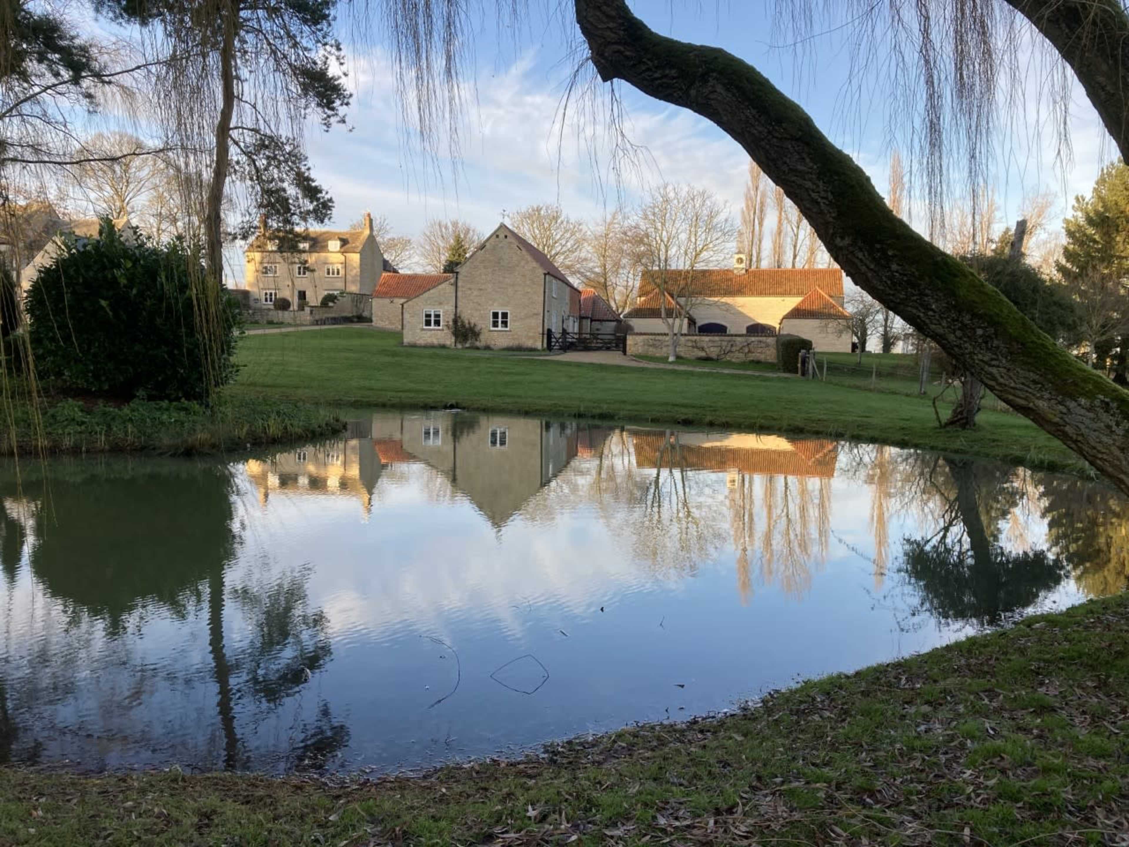 A calm pond reflects buildings and trees in a rural landscape.