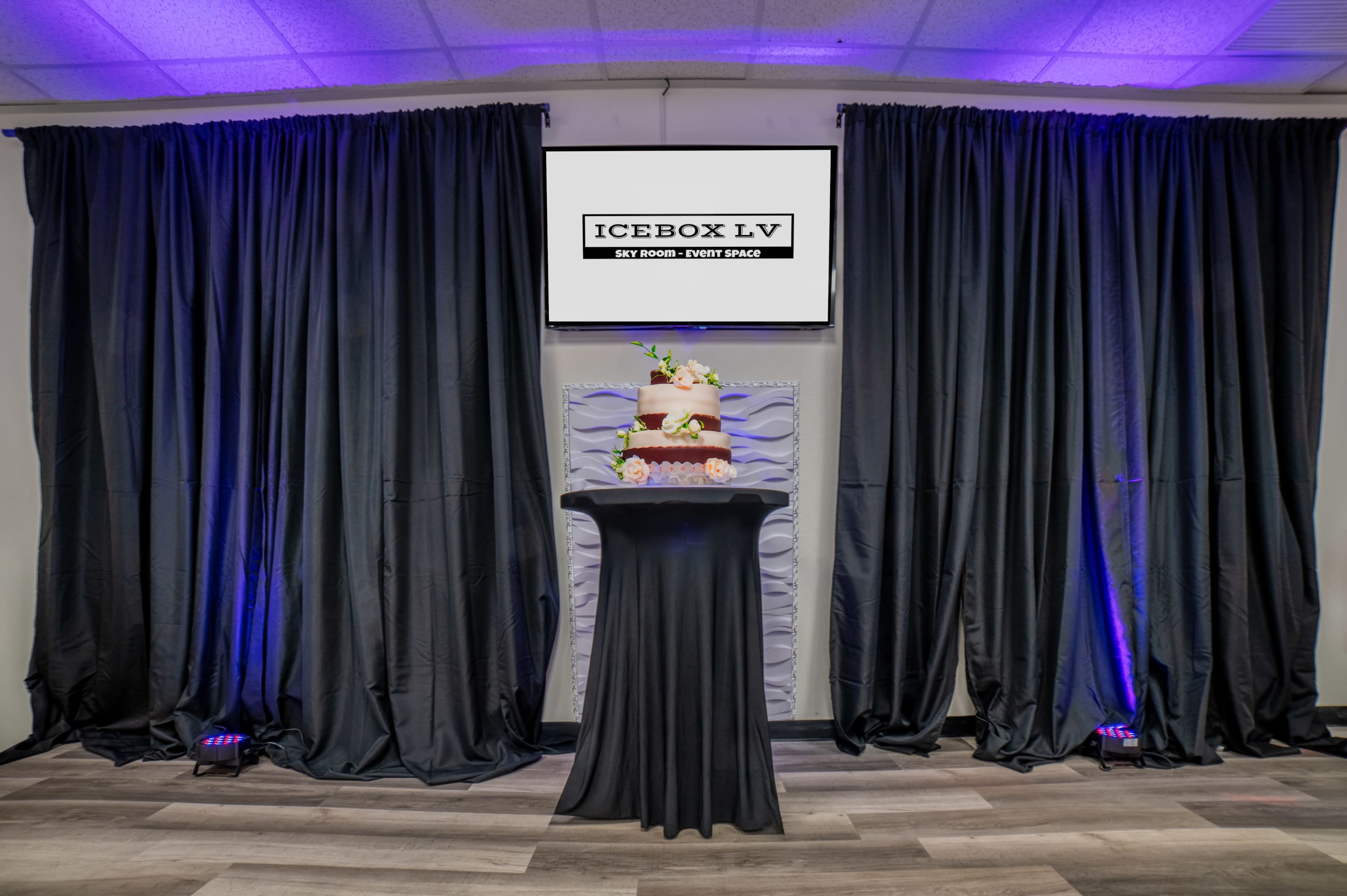 A decorated cake is displayed on a pedestal in front of a large screen and black curtains in an event space.