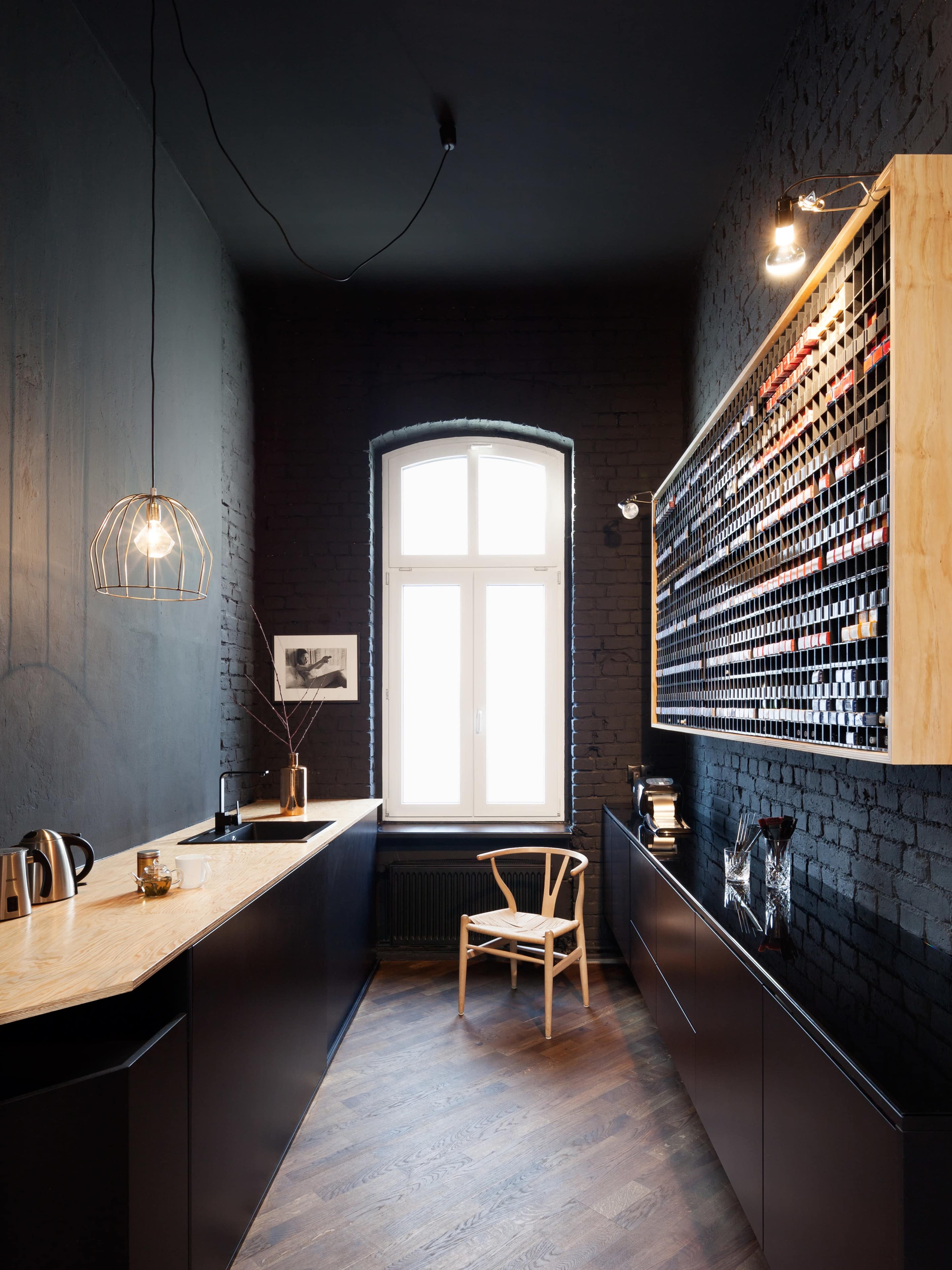 A modern kitchen with dark brick walls, a wooden counter, a wine rack filled with bottles, and a single chair beside a large window.