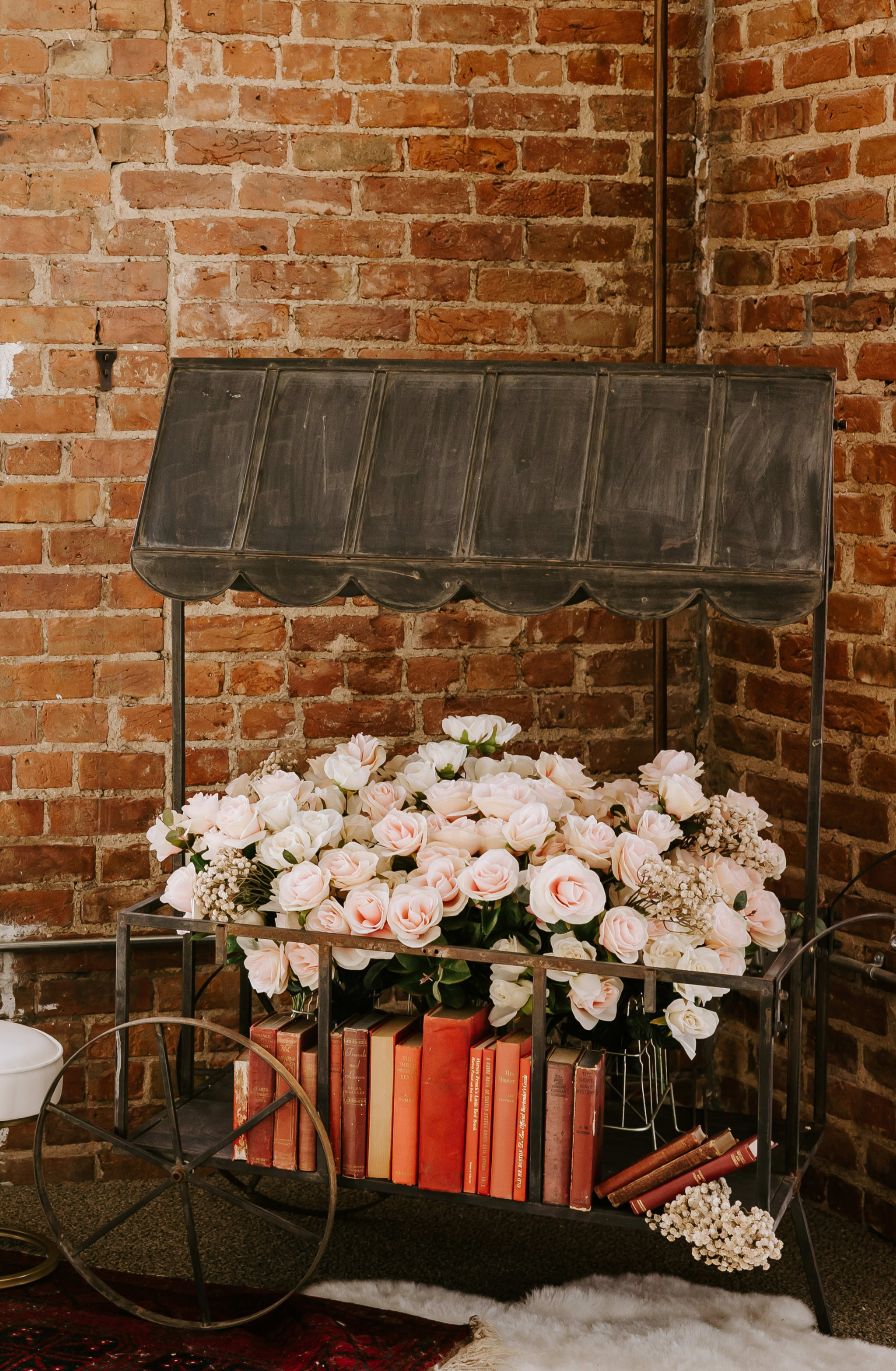 A vintage cart filled with pink roses and stacked with books stands against a brick wall.
