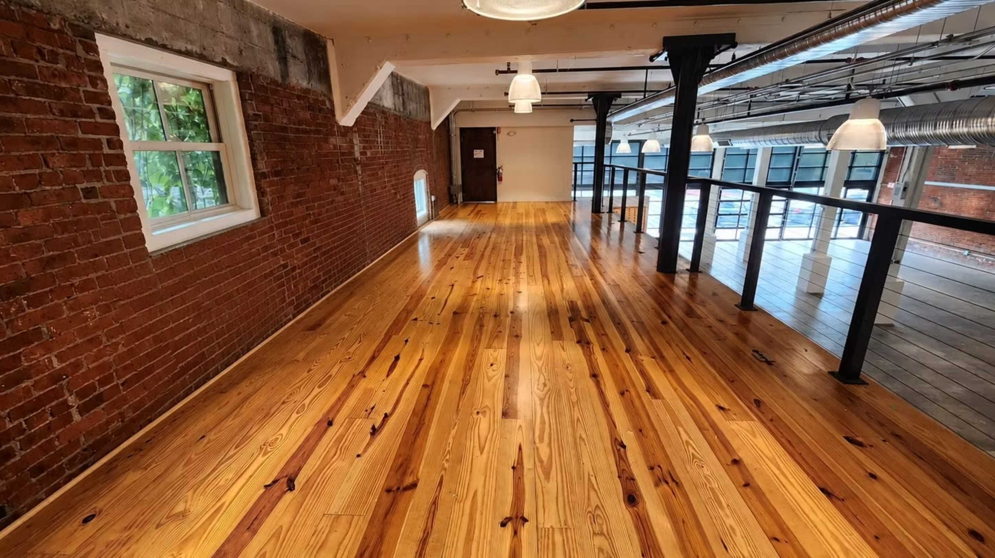 The image shows a spacious interior with polished wooden floors, exposed brick walls, and large windows.