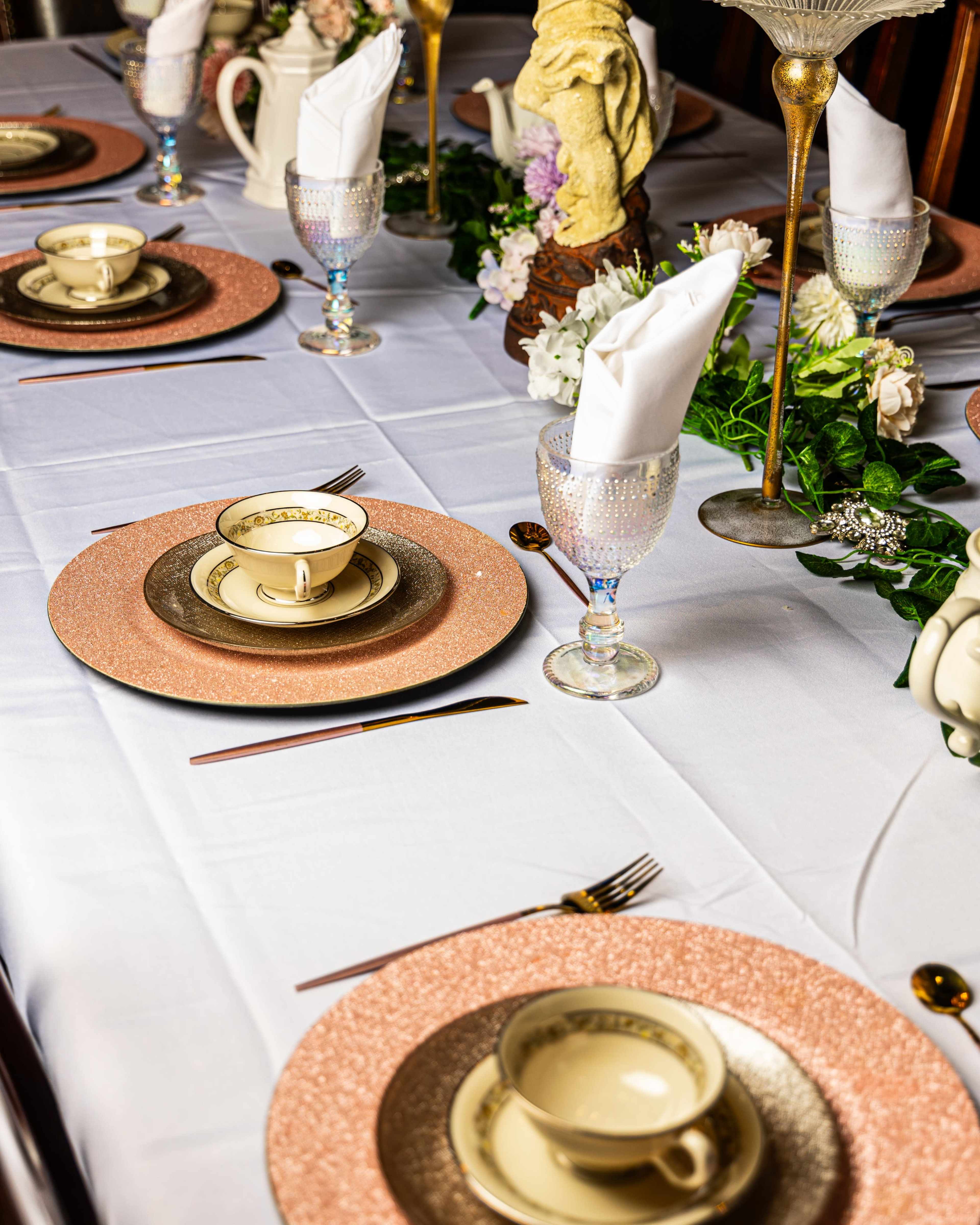 A formal dining table is set with elegant plates, cups, and decorative glassware, surrounded by flowers and a centerpiece.