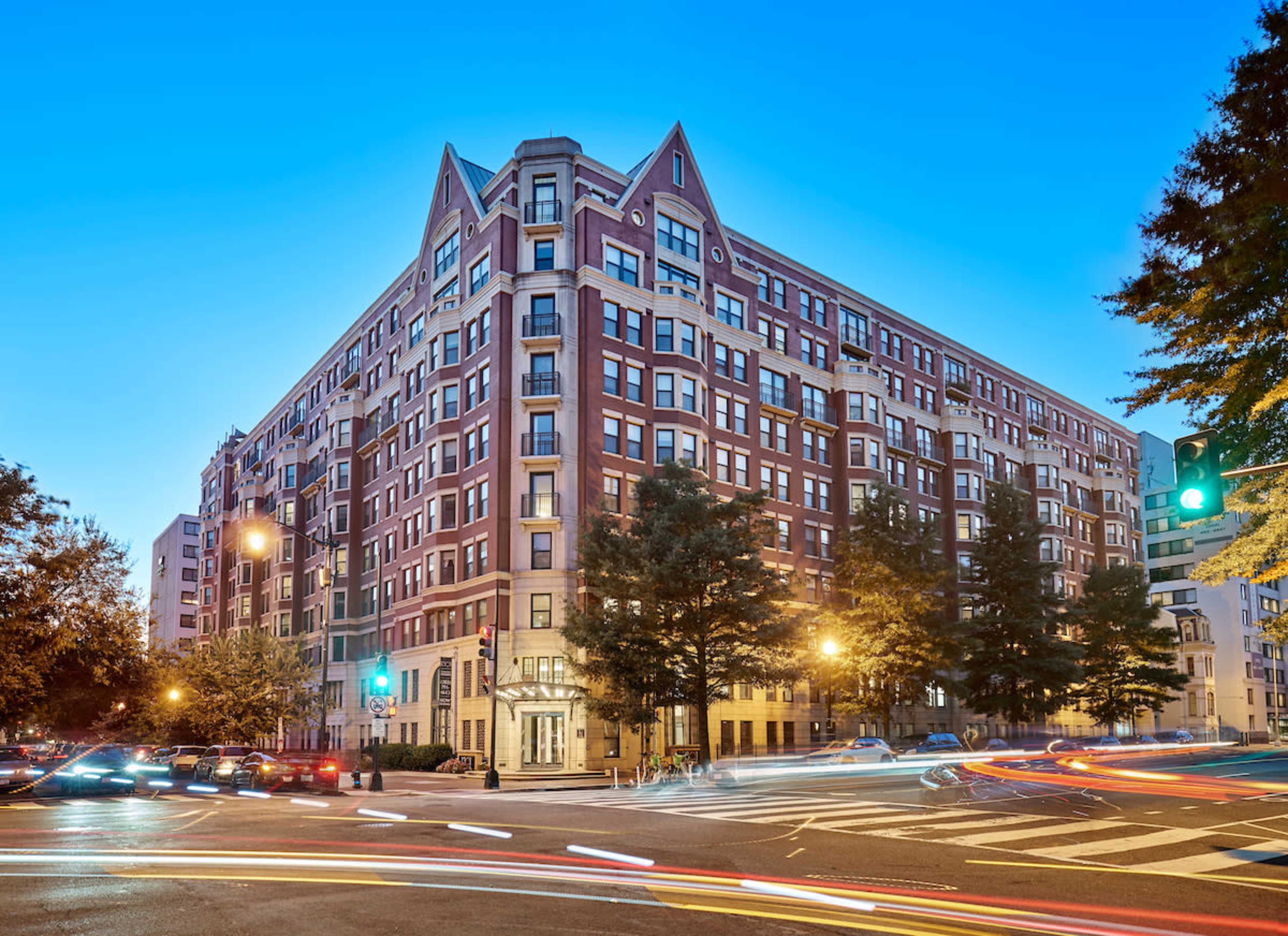A large, brick apartment building with a peaked roof stands at the corner of a city intersection during twilight, with vehicle lights streaking by.