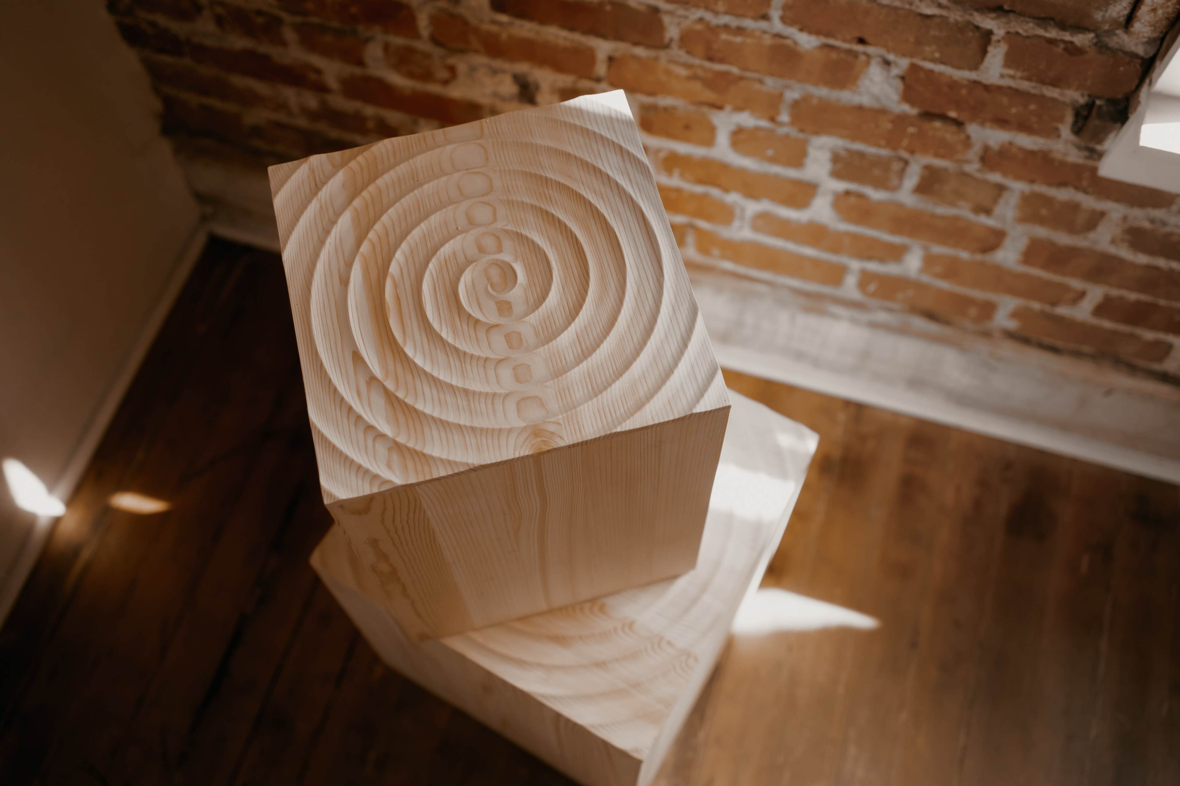 Two wooden cubes stacked on top of each other, with intricate circular patterns carved into their surfaces, against a backdrop of exposed brick walls.