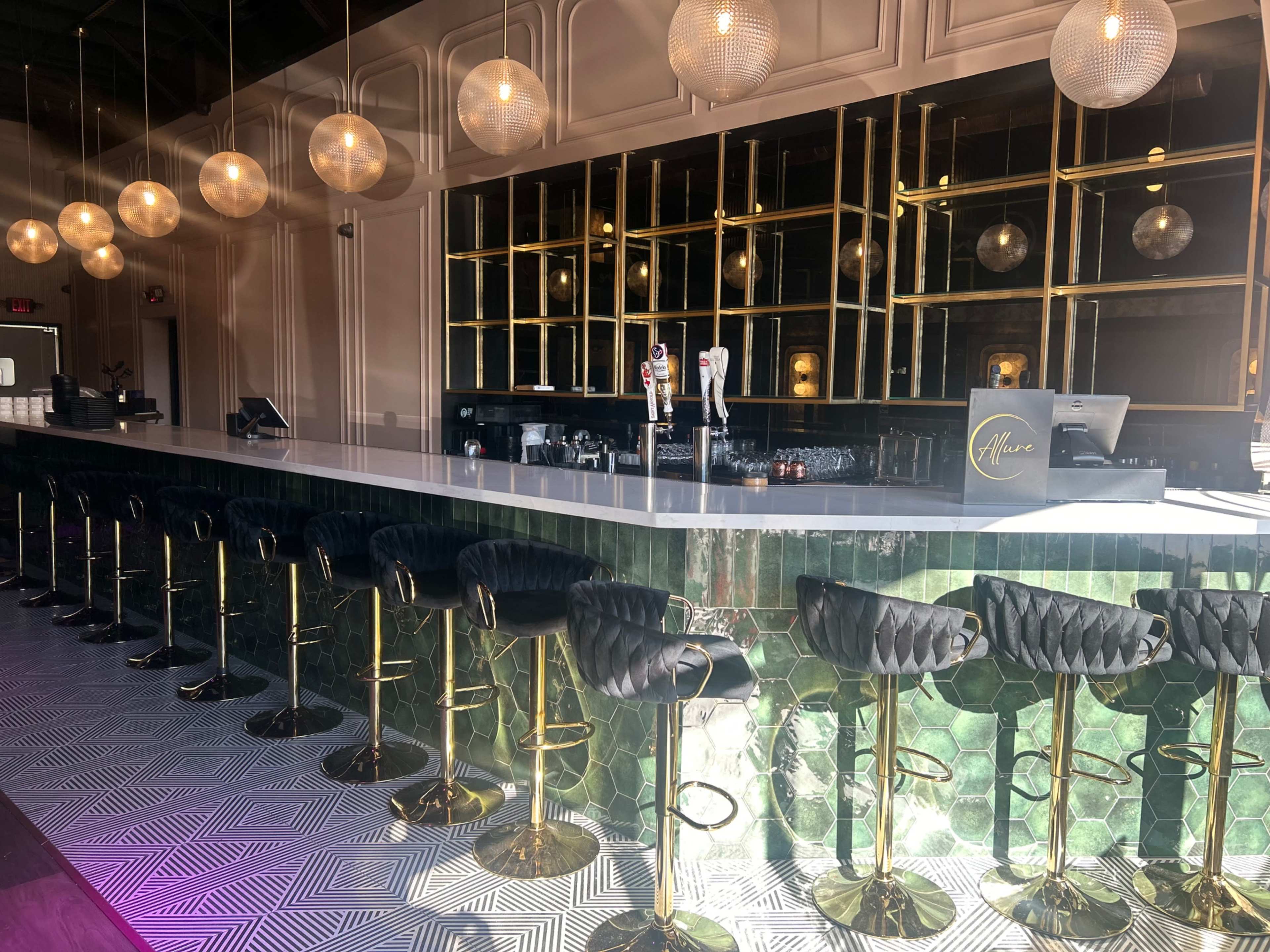 The image shows a modern bar interior with a sleek white countertop, black and gold stools, and decorative lighting fixtures.
