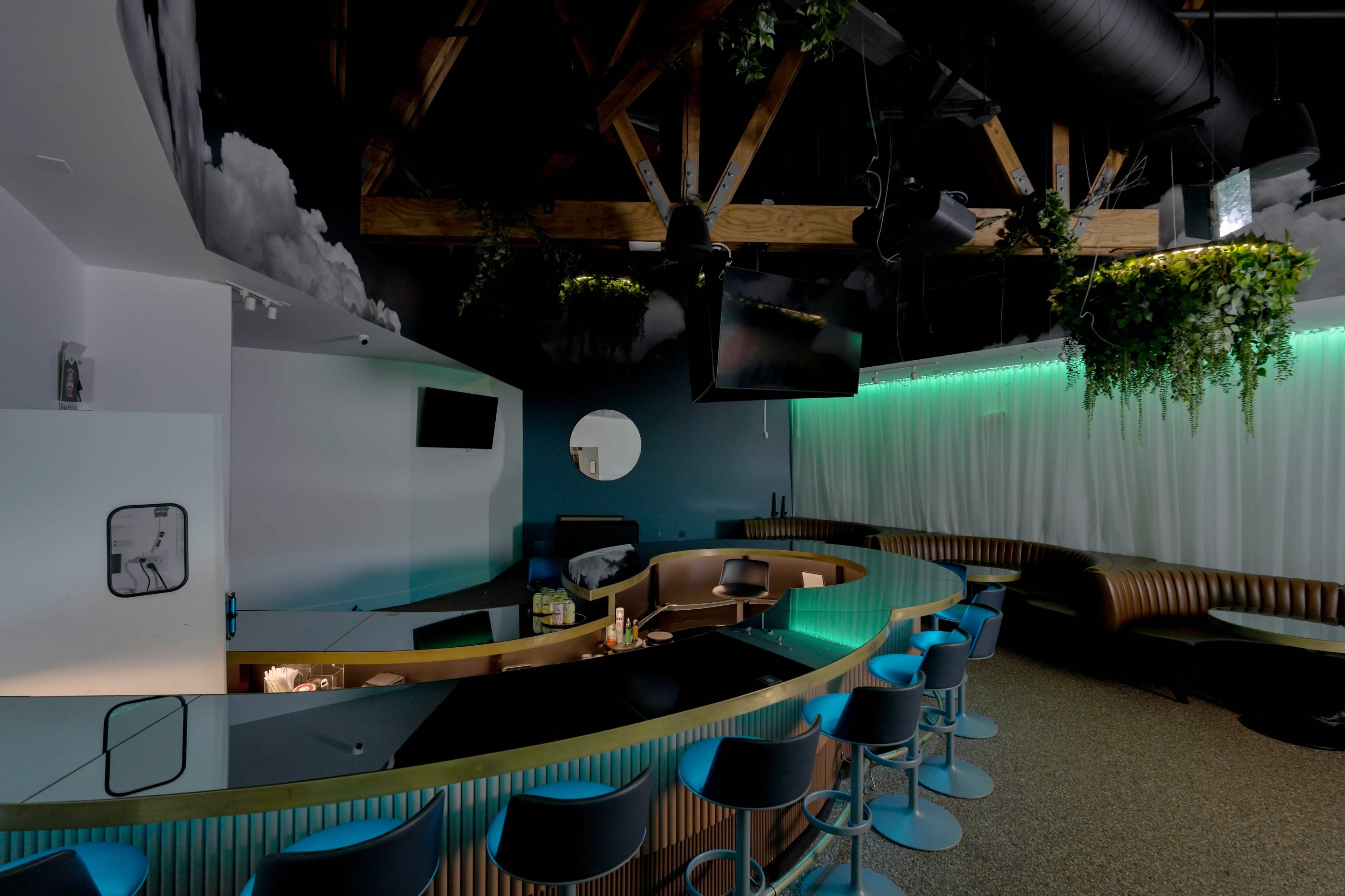 The image shows a modern bar area with a curved counter, high stools, and greenery hanging from the ceiling, illuminated by blue and green lights.