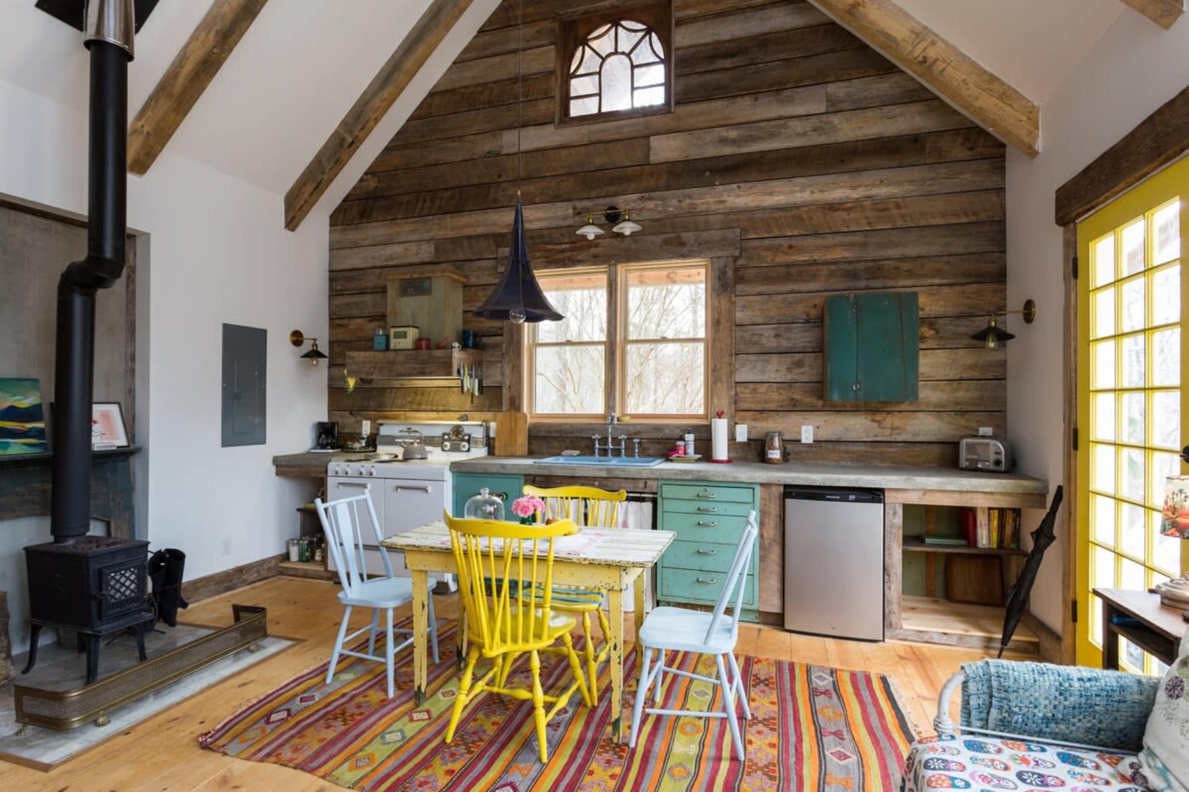 A rustic kitchen features a wooden dining table with colorful chairs, a stove, and a large window overlooking the outdoors.