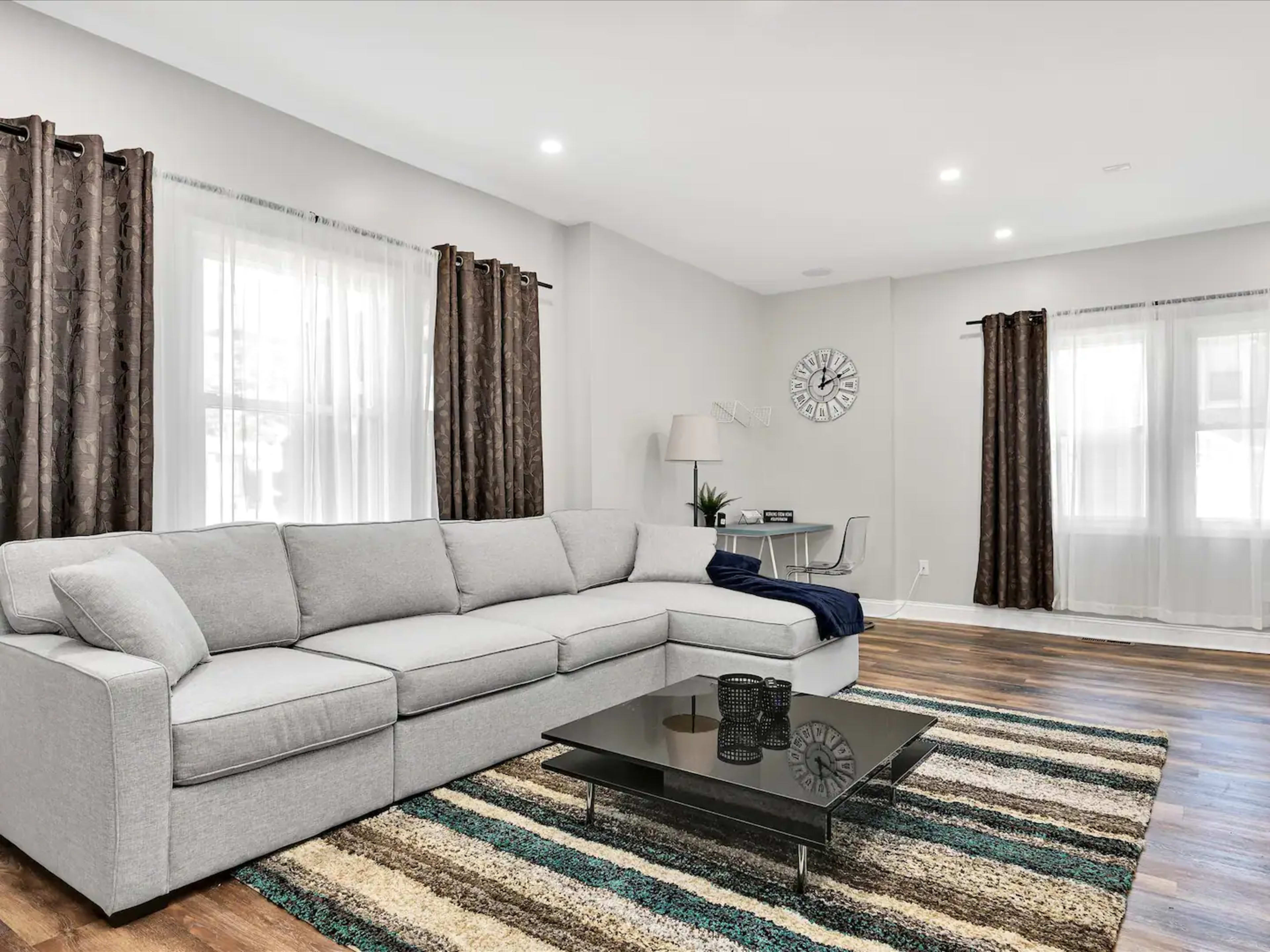 A modern living room features a gray sectional sofa, a glass coffee table on a striped rug, and windows with brown curtains.