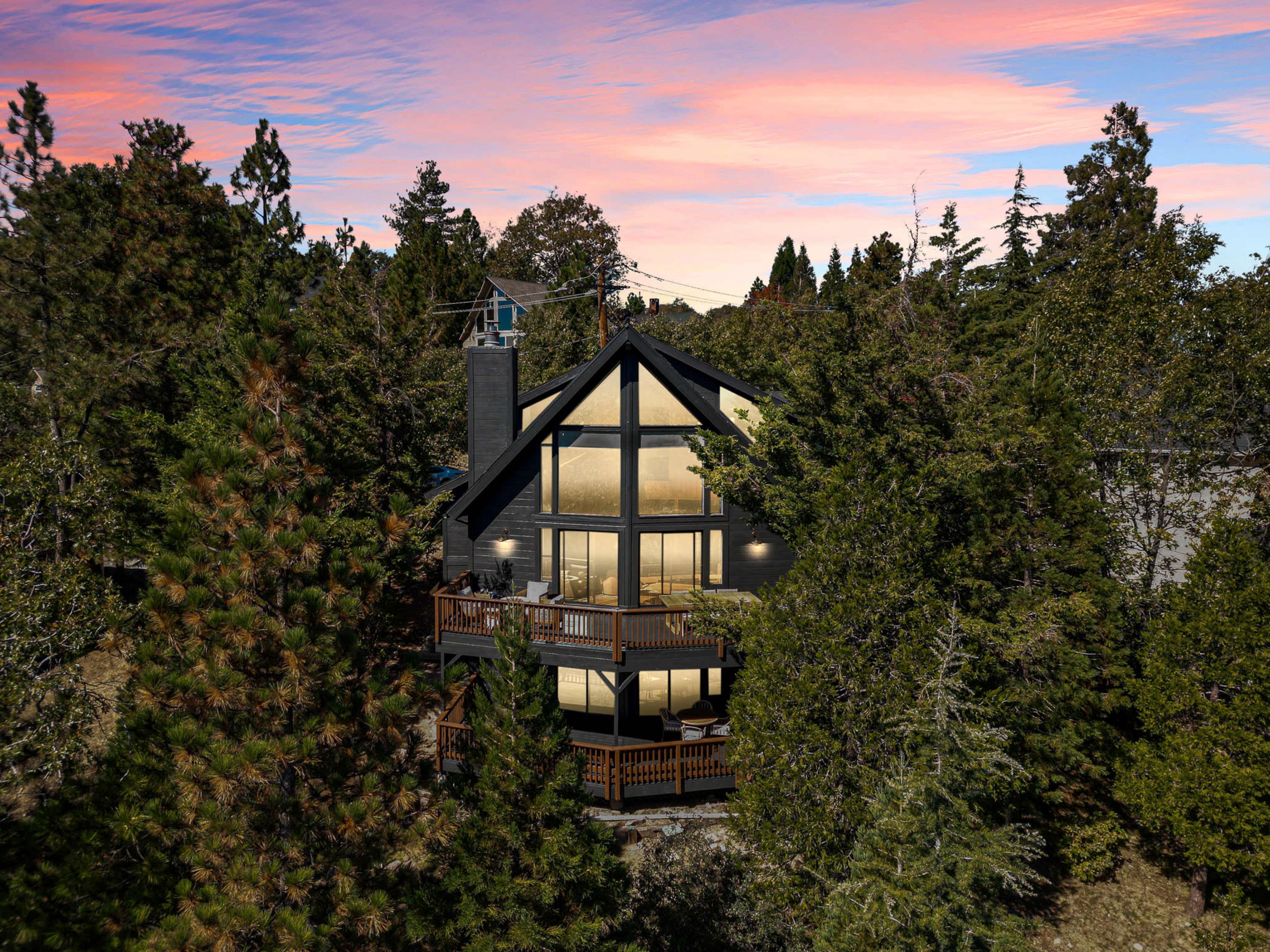 A two-story black A-frame house is surrounded by tall trees under a colorful sunset sky.