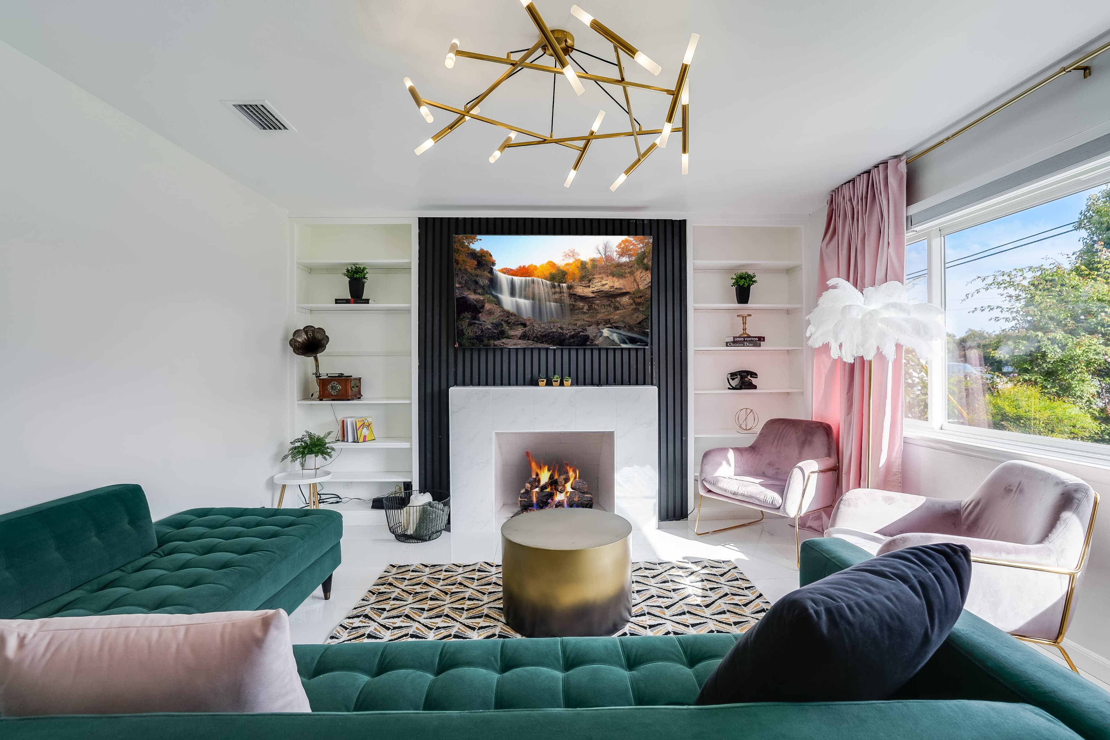 A modern living room features green velvet sofas, a round gold coffee table, a fireplace, and a large television displaying a waterfall scene.