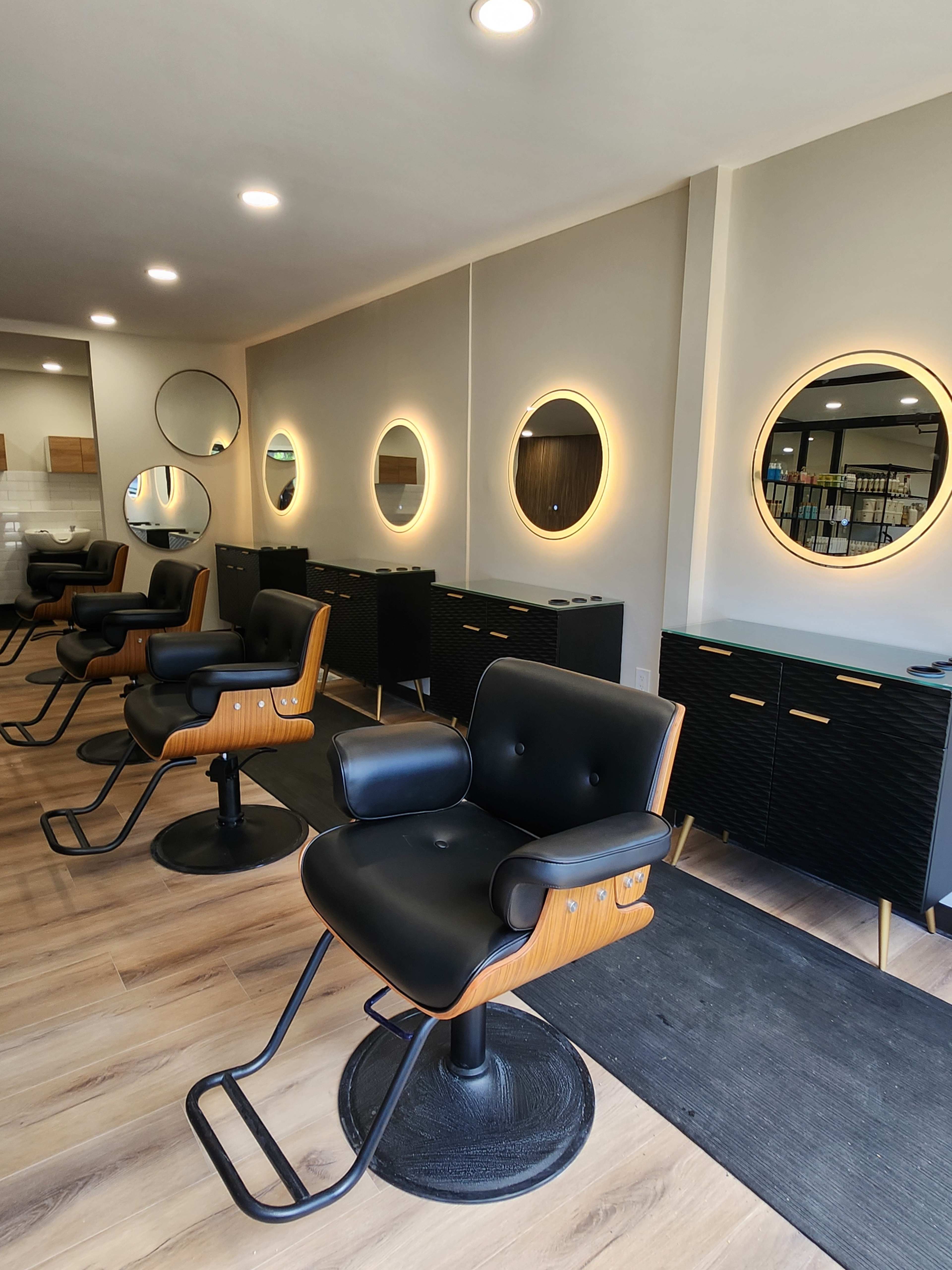 A modern hair salon features black styling chairs arranged in front of illuminated round mirrors and sleek dark cabinetry.