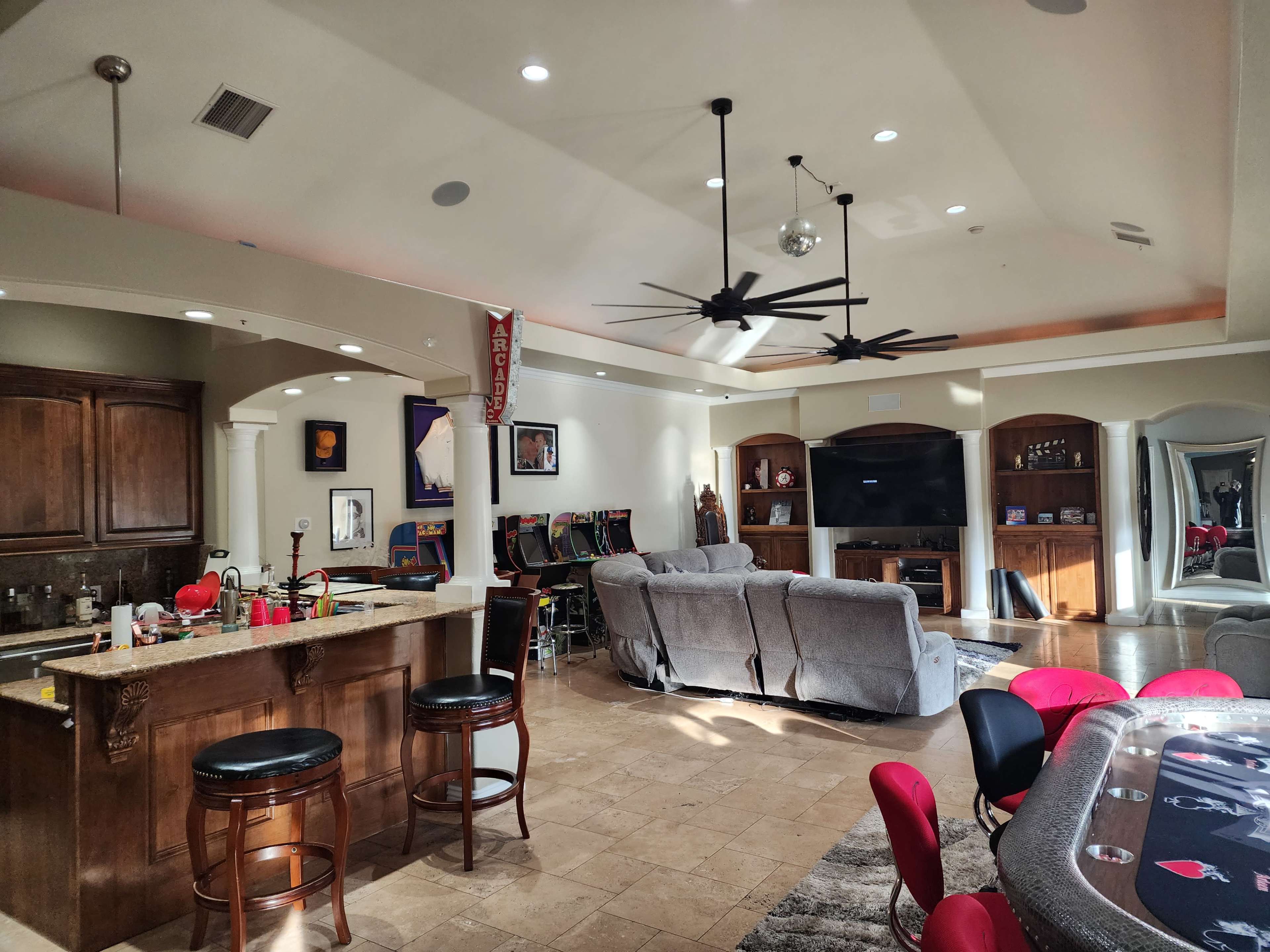 A spacious living area featuring a bar with stools, a poker table with red chairs, and a seating area with gray sofas.