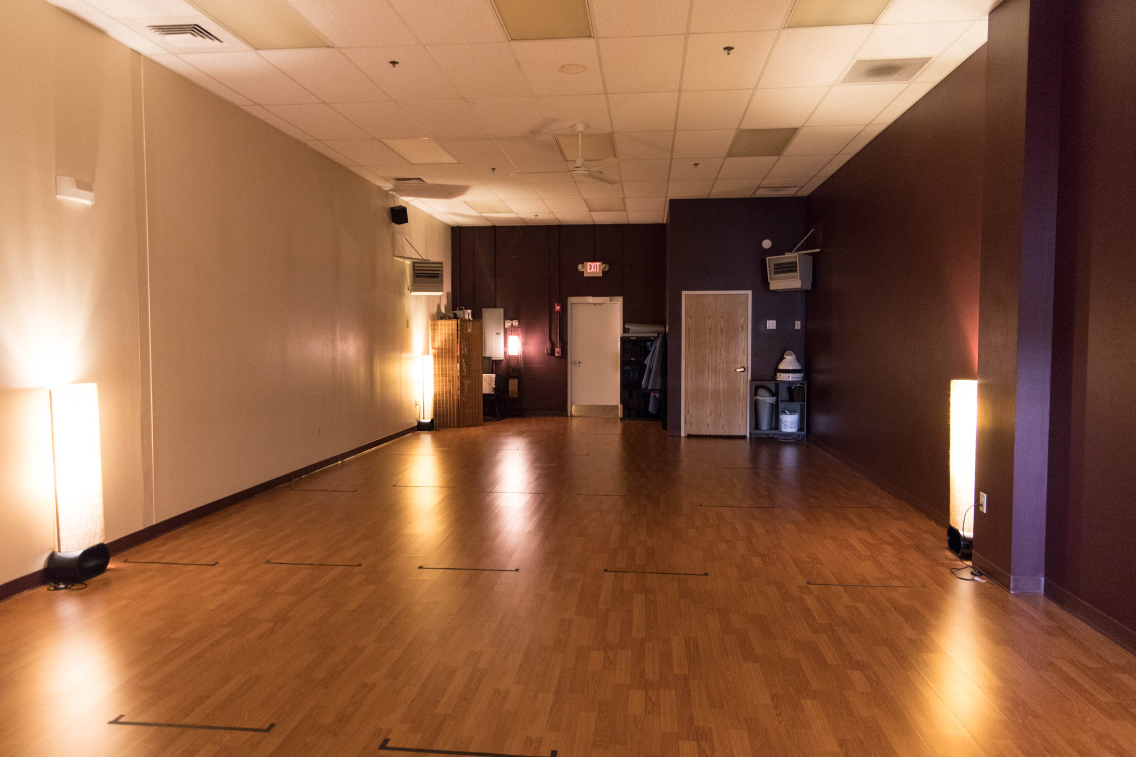 An empty, well-lit studio space with wooden flooring, featuring two tall lamps on either side and a door leading to another room.