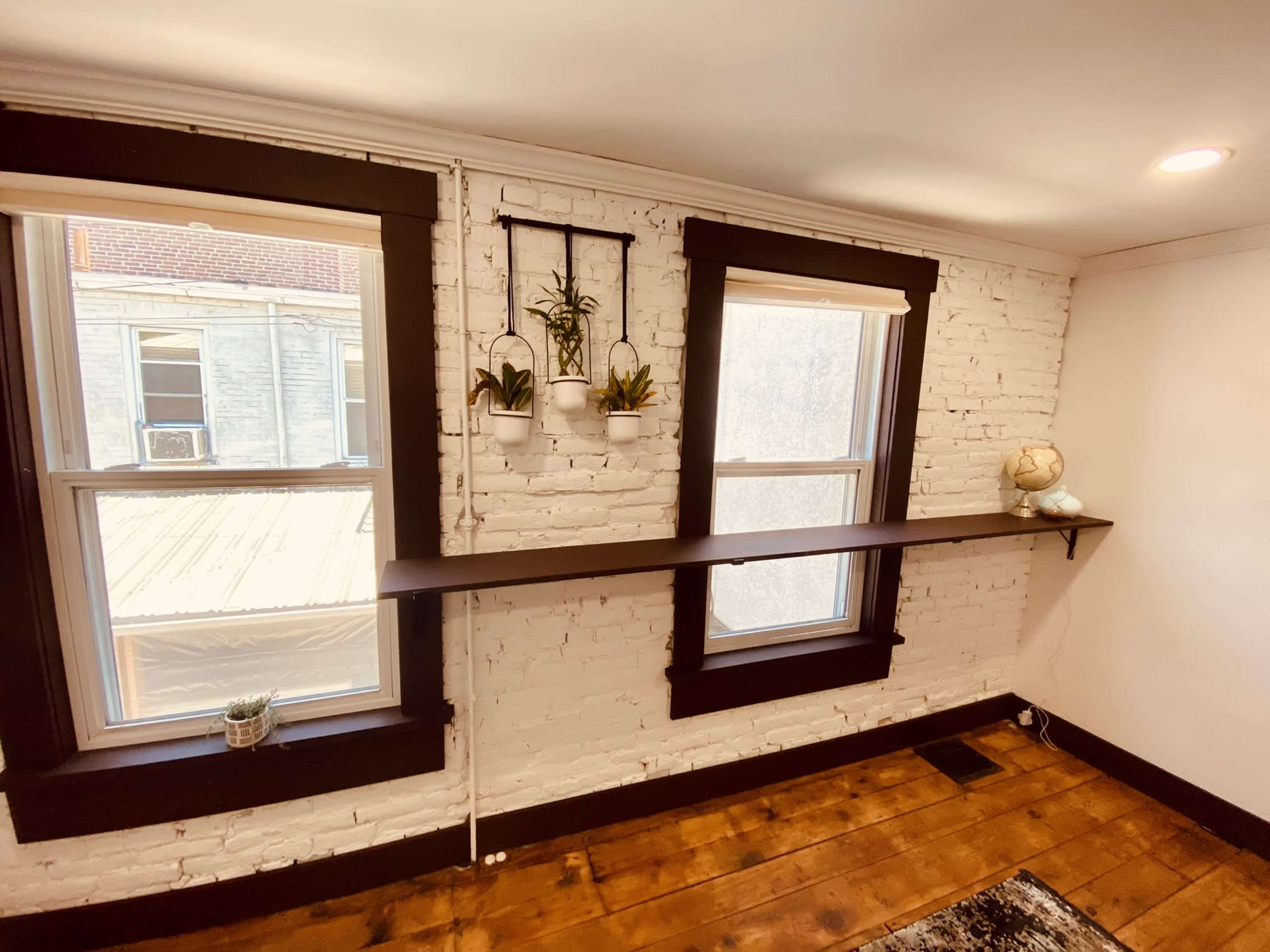 A room with two windows and a horizontal shelf mounted on a white brick wall, featuring small potted plants and a globe.