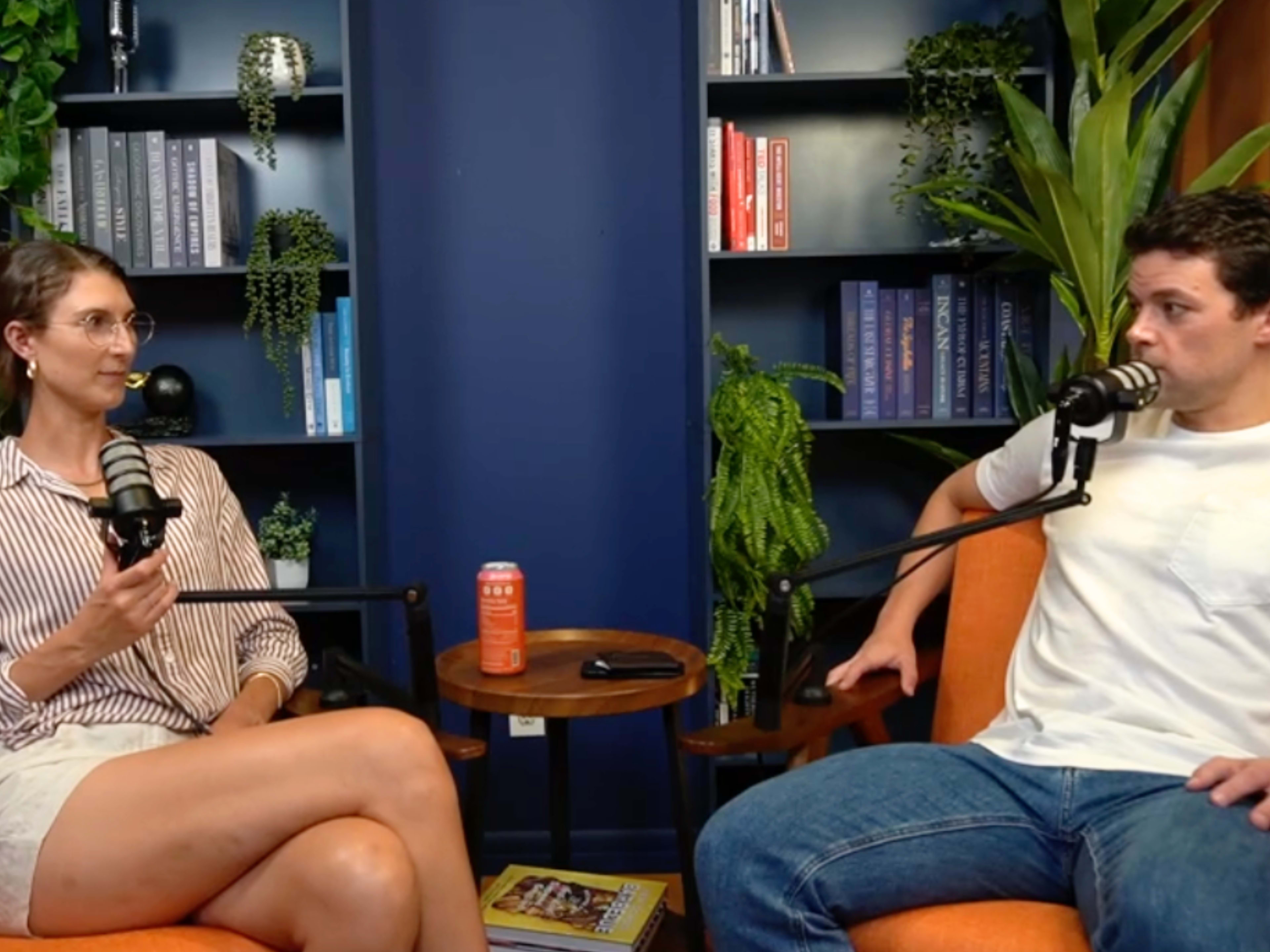 Two individuals are seated in a well-decorated room with bookshelves and plants, both holding microphones and engaged in conversation.