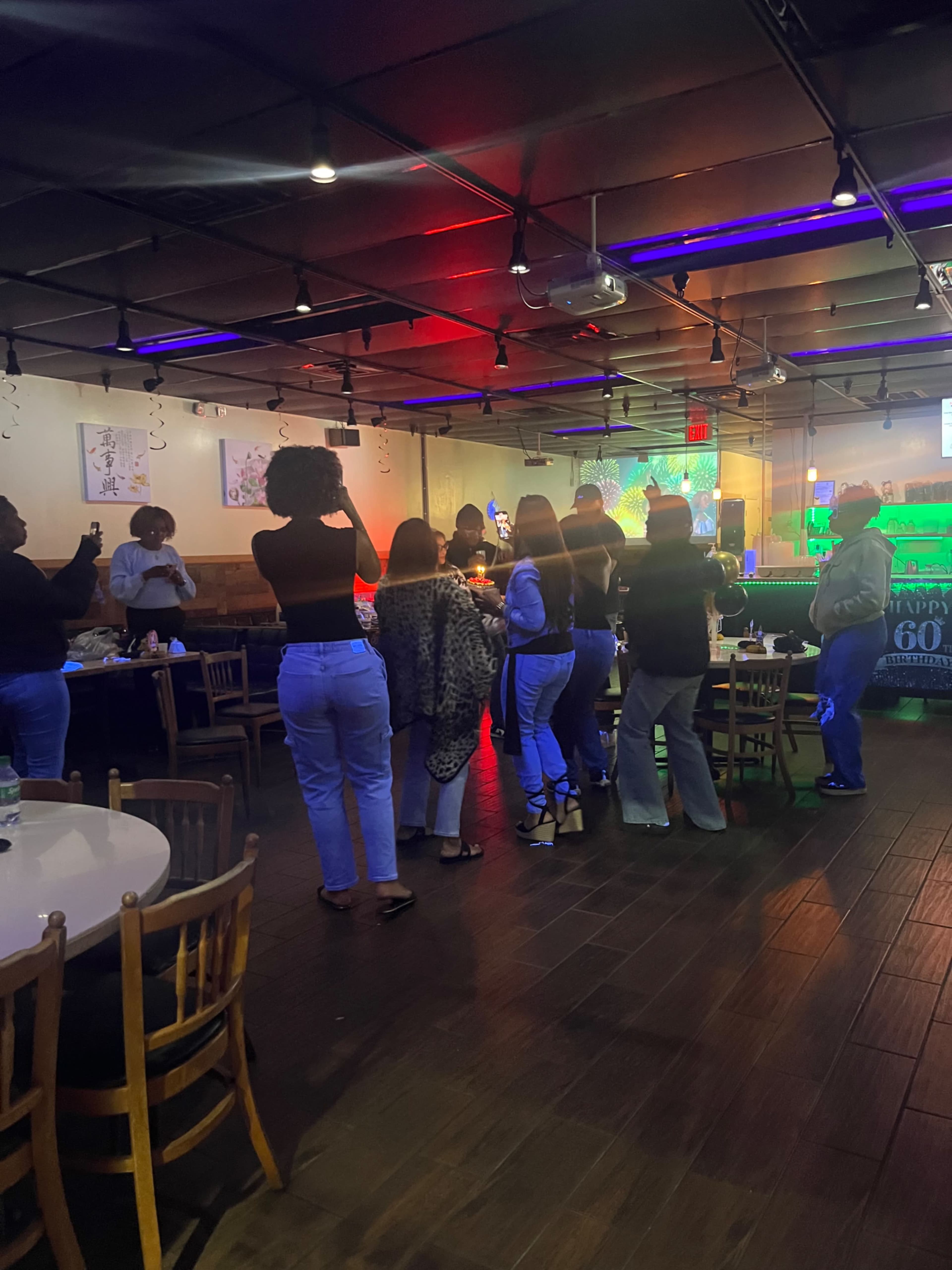 A group of people stands and dances in a lively restaurant, with colorful lights illuminating the space.