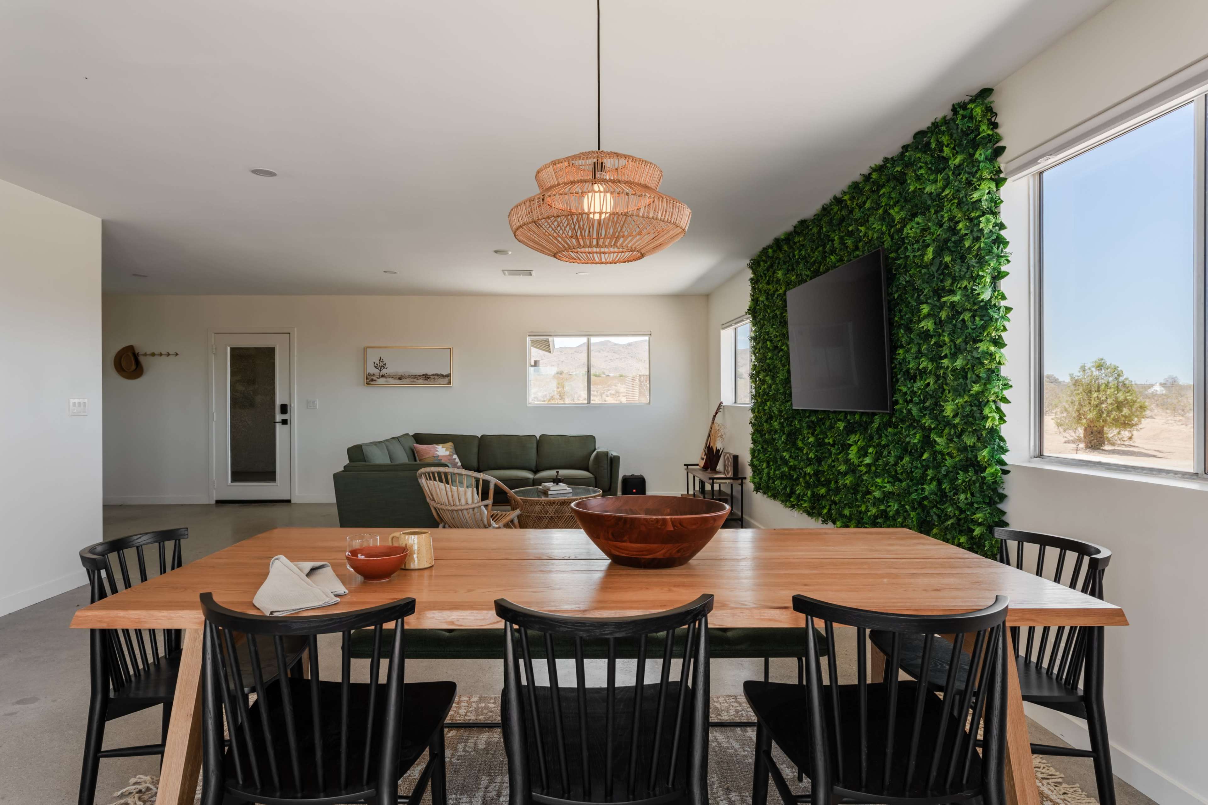 The image shows a modern dining area with a wooden table and black chairs, adjacent to a living space featuring a green sofa and a wall of artificial greenery.