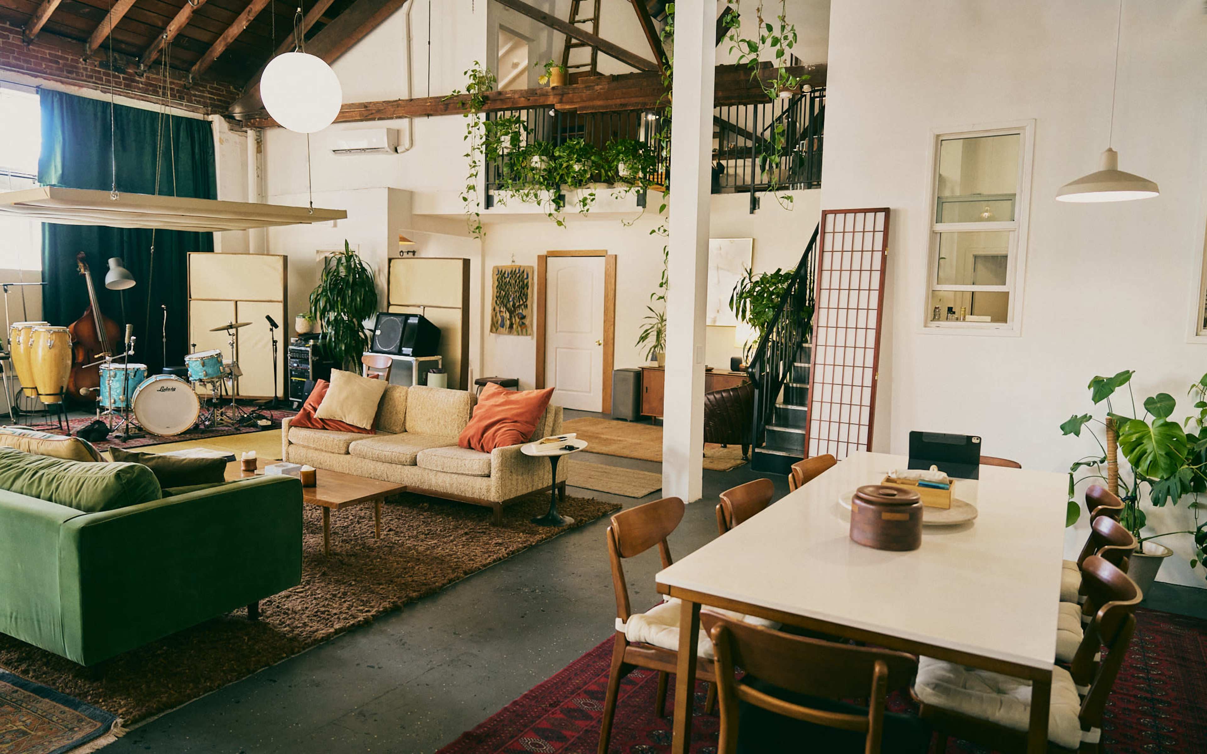 The image shows an open-concept living space with a mix of furniture, including a sofa, dining table, and musical instruments set up in a room adorned with plants and large windows.