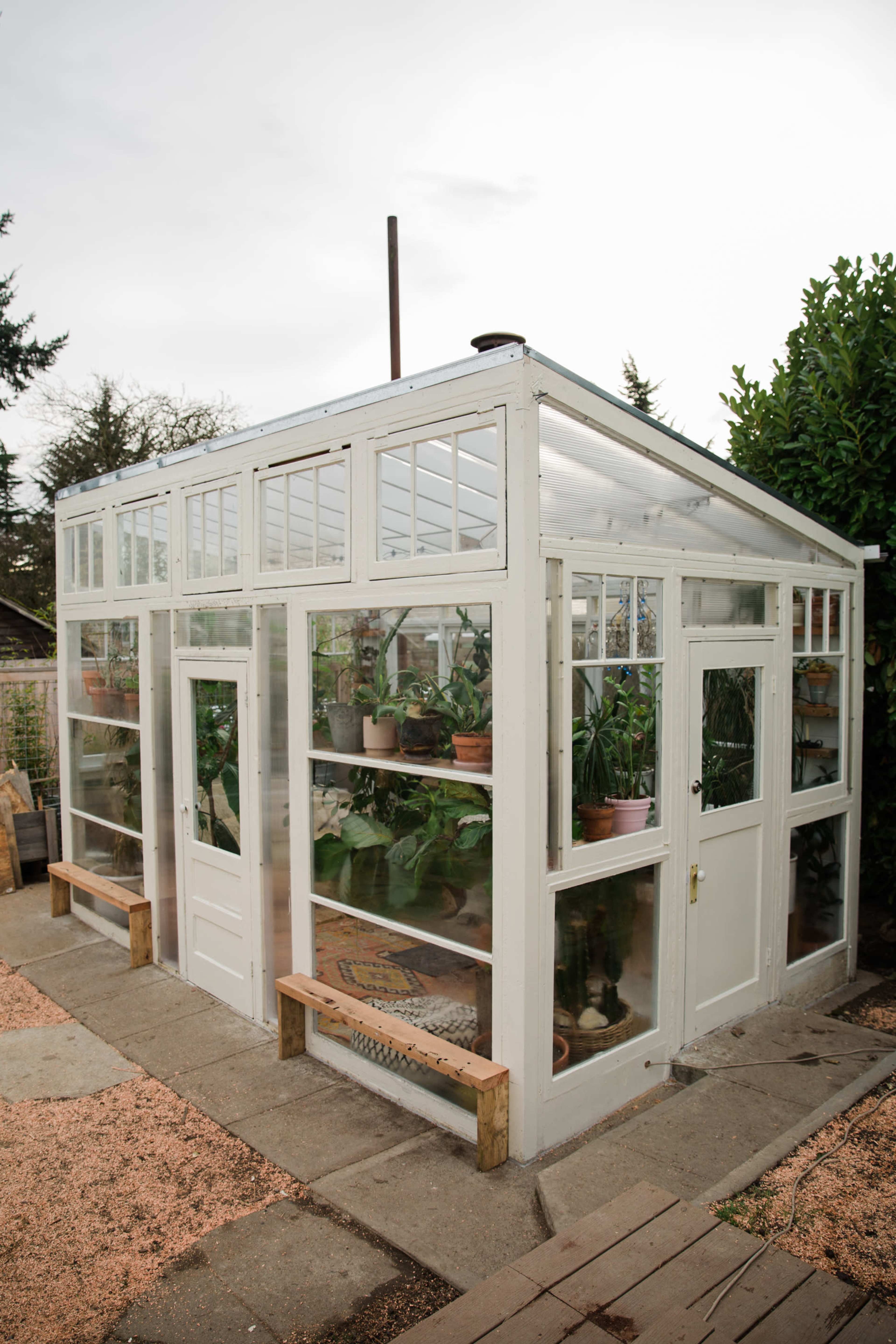 A white greenhouse with glass panels houses various potted plants on shelves inside.