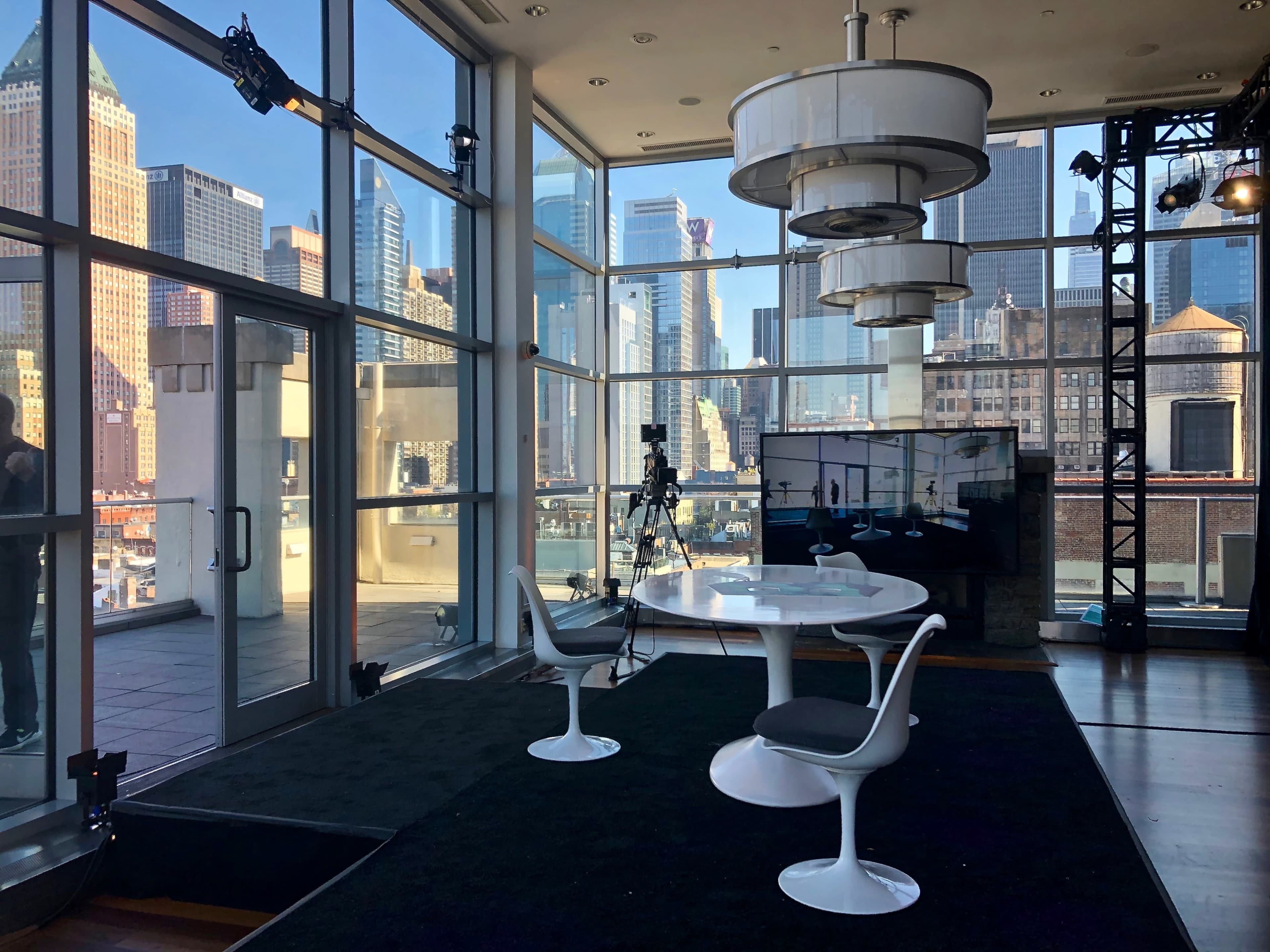 A modern studio setup with a round table and chairs overlooks a city skyline through large glass windows.