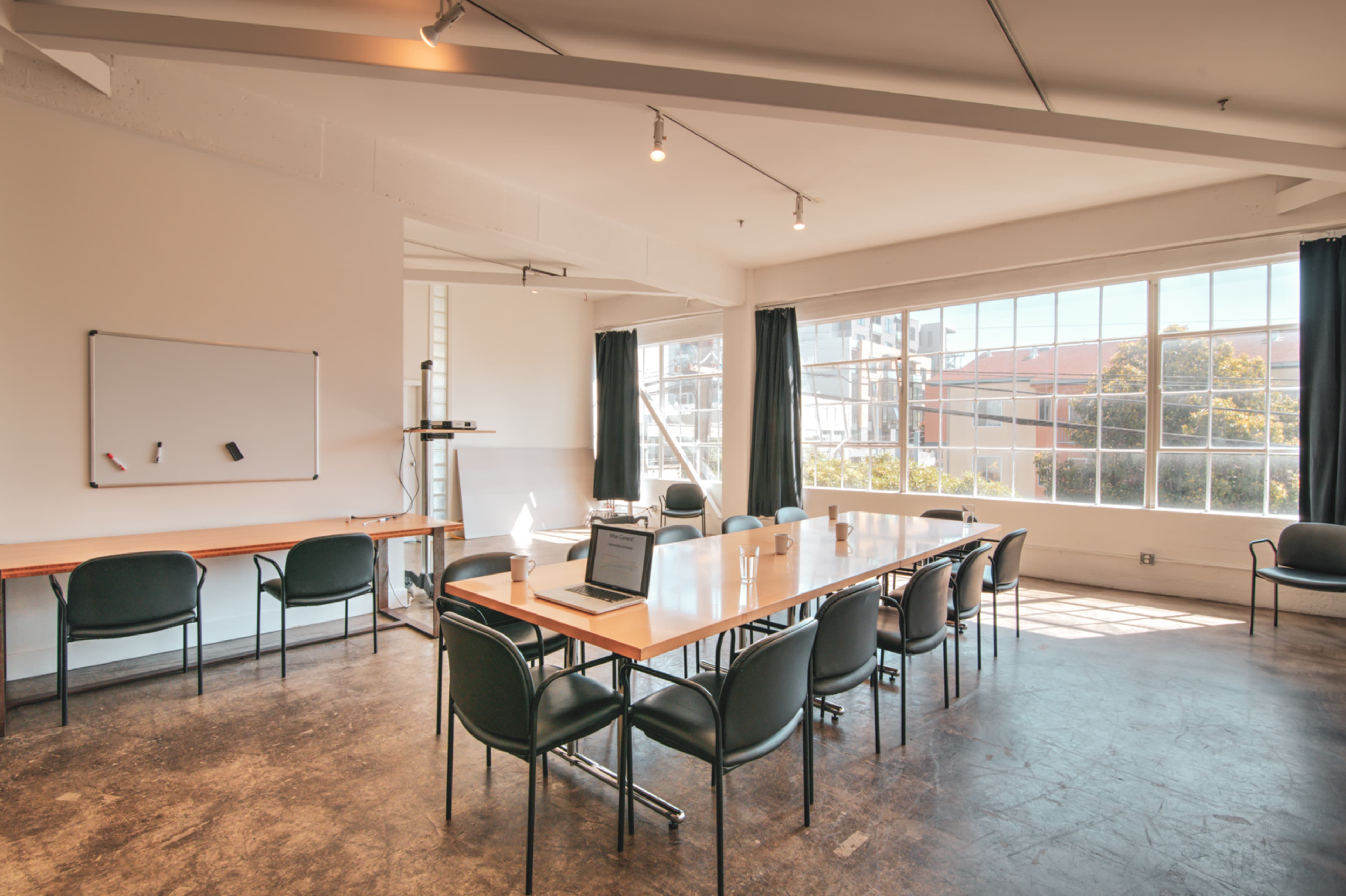 A bright conference room features a large table surrounded by black chairs, with a whiteboard on one wall and large windows offering a view outside.