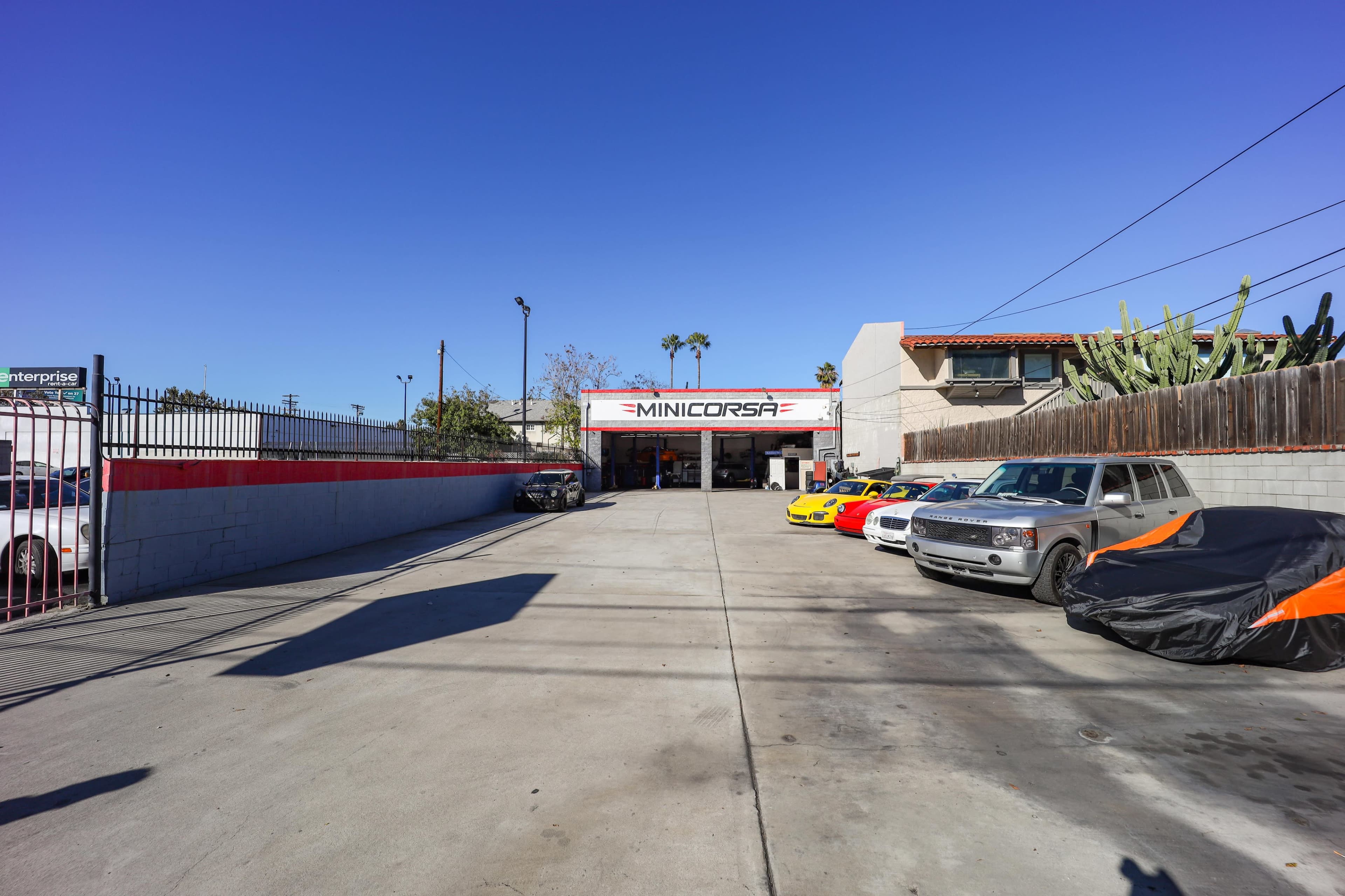 A clear view of a car workshop named "MINICORSA," featuring several cars parked in front and a bright blue sky overhead.