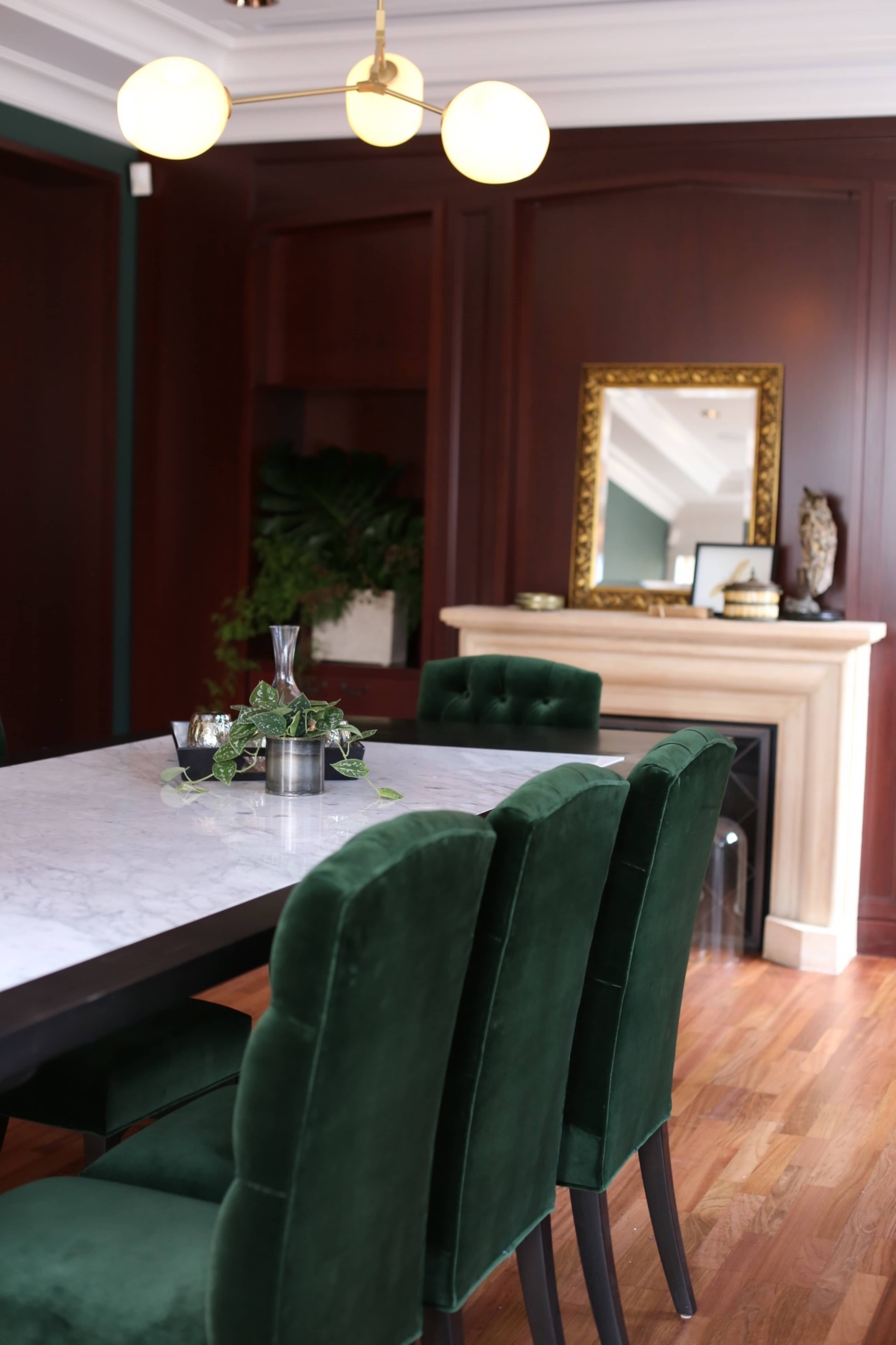 A dining area features a marble table surrounded by green upholstered chairs, with a decorative fireplace and a gold-framed mirror in the background.