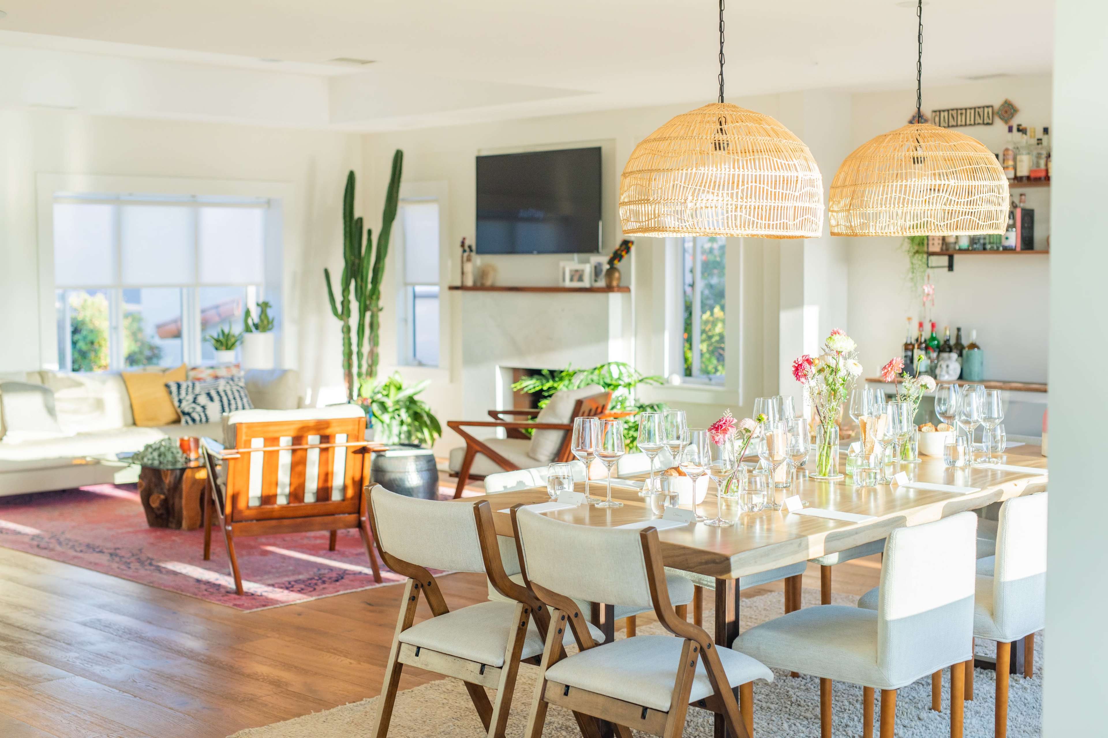 A bright, airy dining room with a wooden table set for a meal, surrounded by light-colored chairs, and decorated with flowers, while a cozy living area is visible in the background.