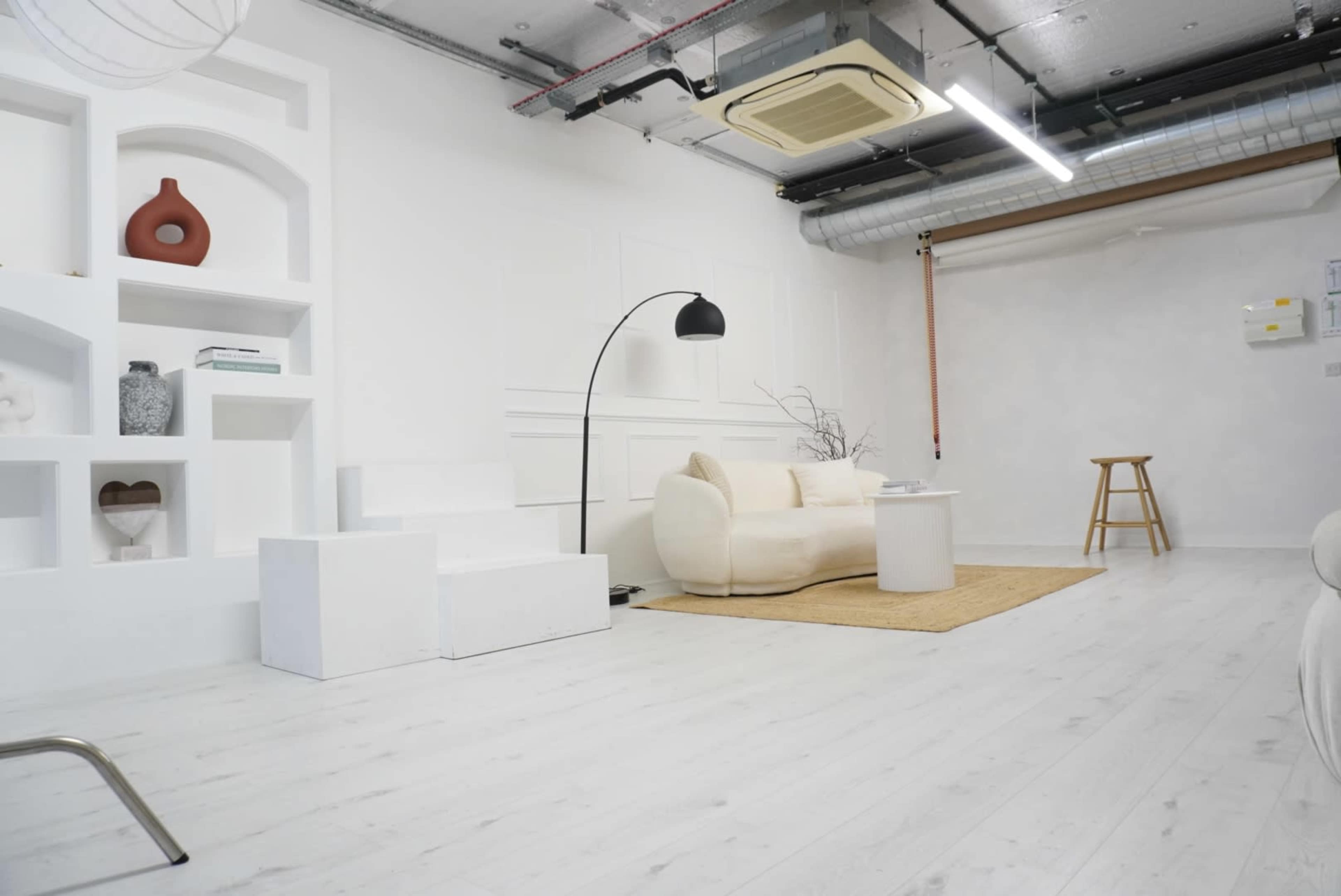 The image shows a minimalistic indoor space featuring a light-colored sofa, a small round table, a rug, and decorative shelves against white walls.