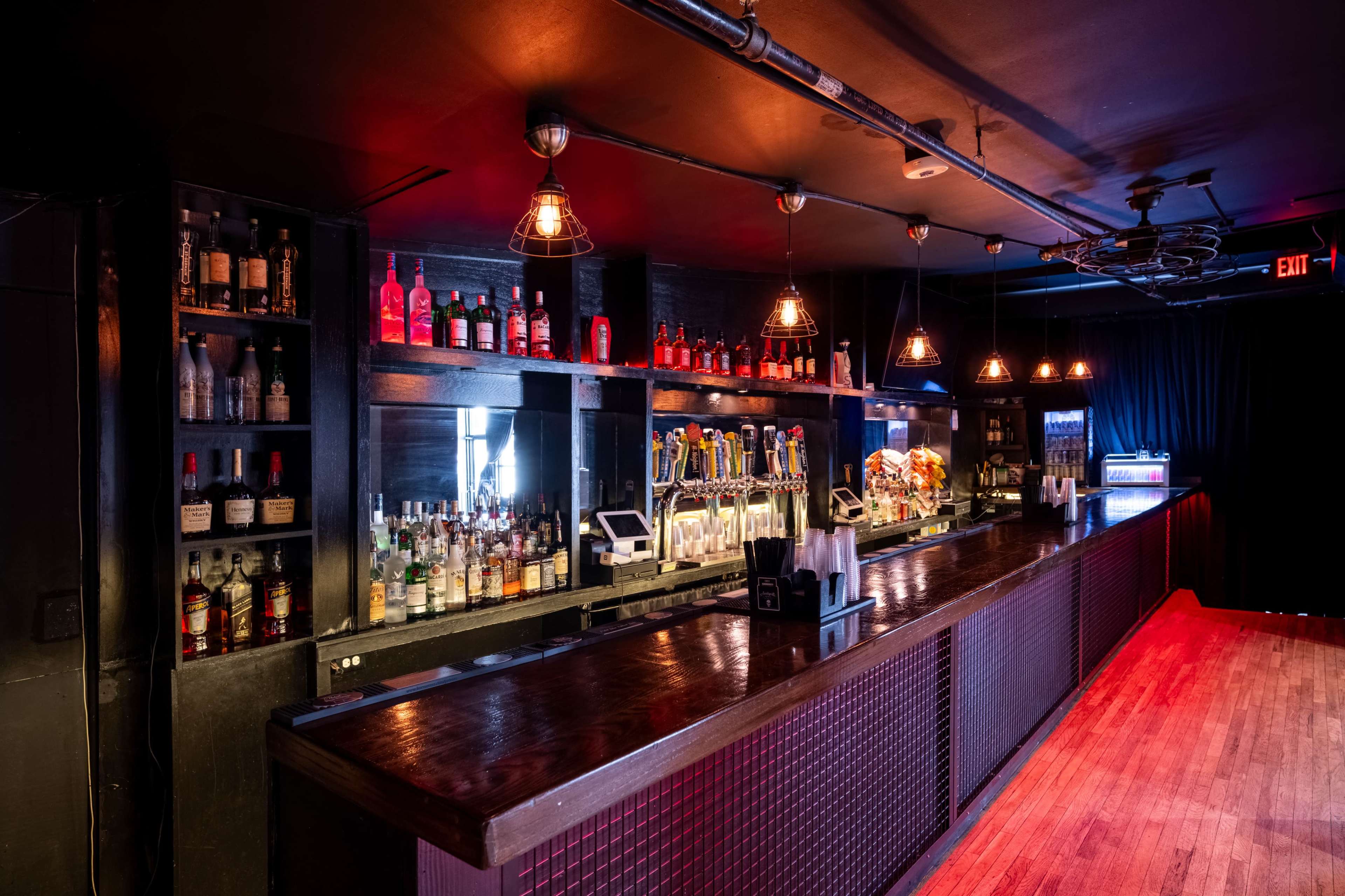 The image shows a dimly lit bar with a wooden counter, shelves filled with various bottles, and illuminated pendant lights hanging overhead.
