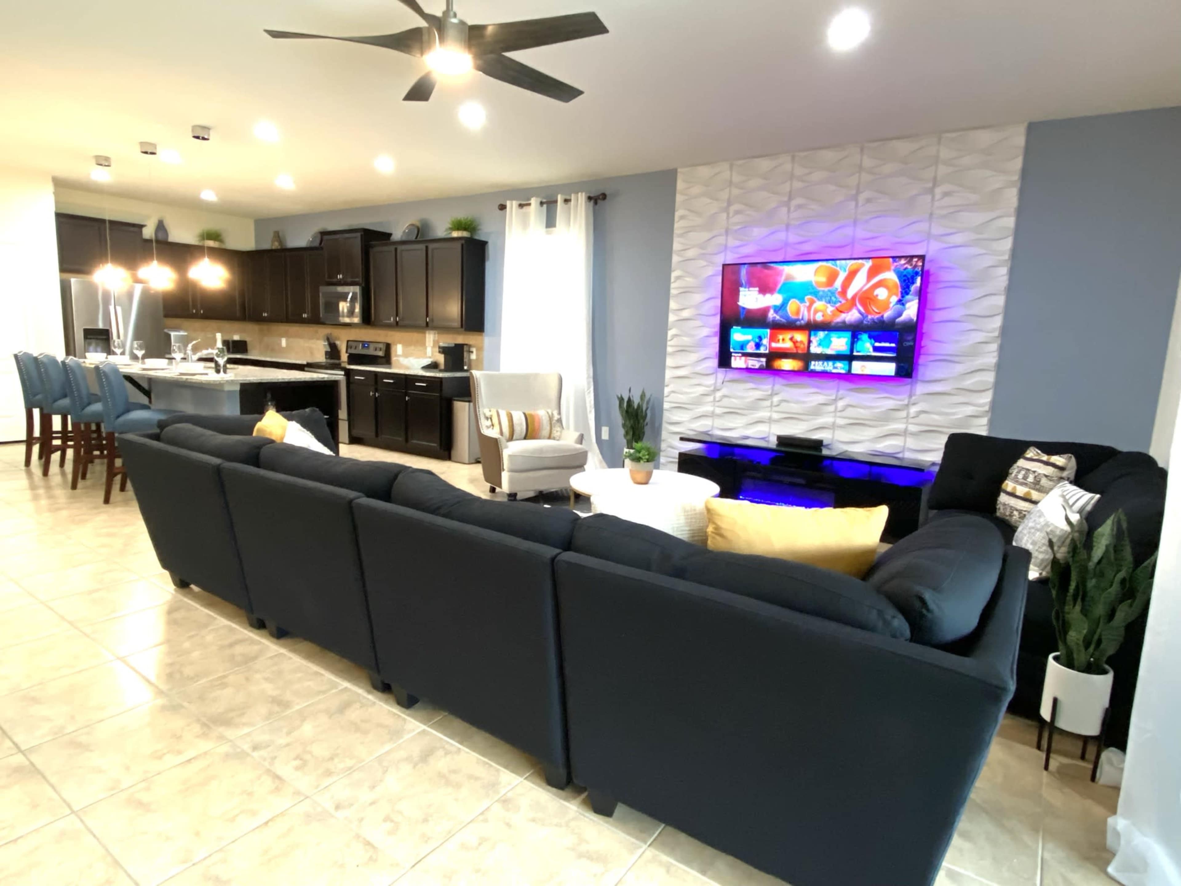The image shows a modern living room with a sectional sofa facing a wall-mounted television, adjacent to a kitchen with dark cabinetry and bar seating.