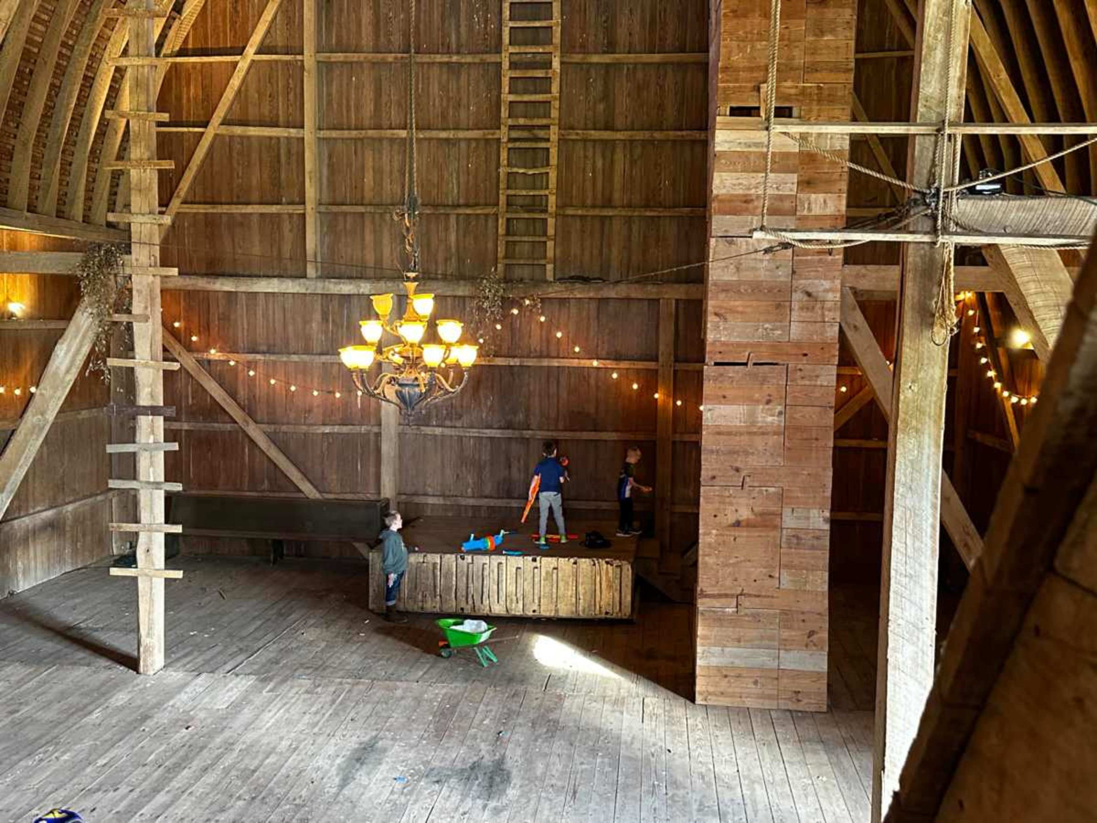The image shows an interior of a rustic barn with wooden beams, a chandelier, and children playing with toys on a wooden platform.