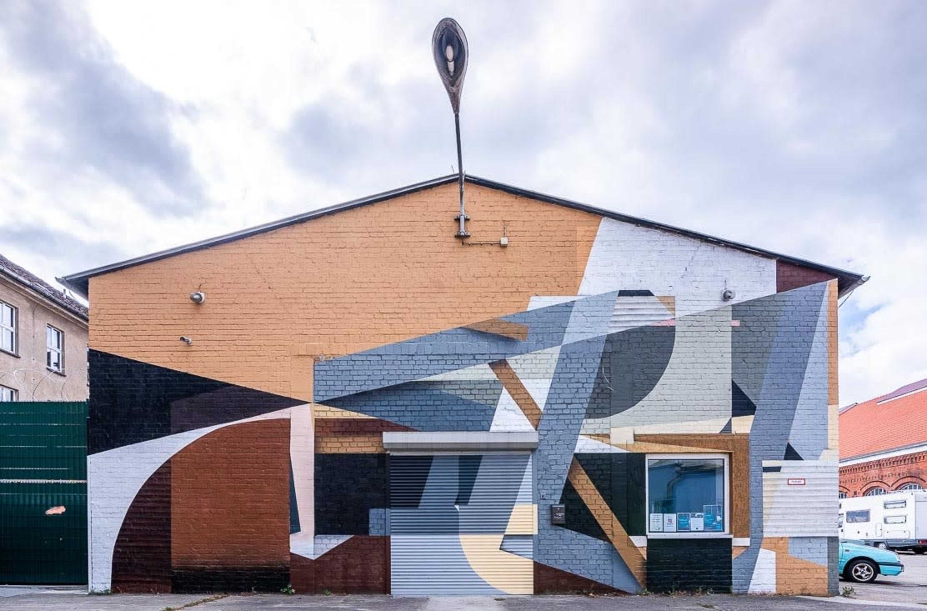 A warehouse building with a large mural featuring abstract geometric shapes in various colors and a large spoon sculpture mounted on top.