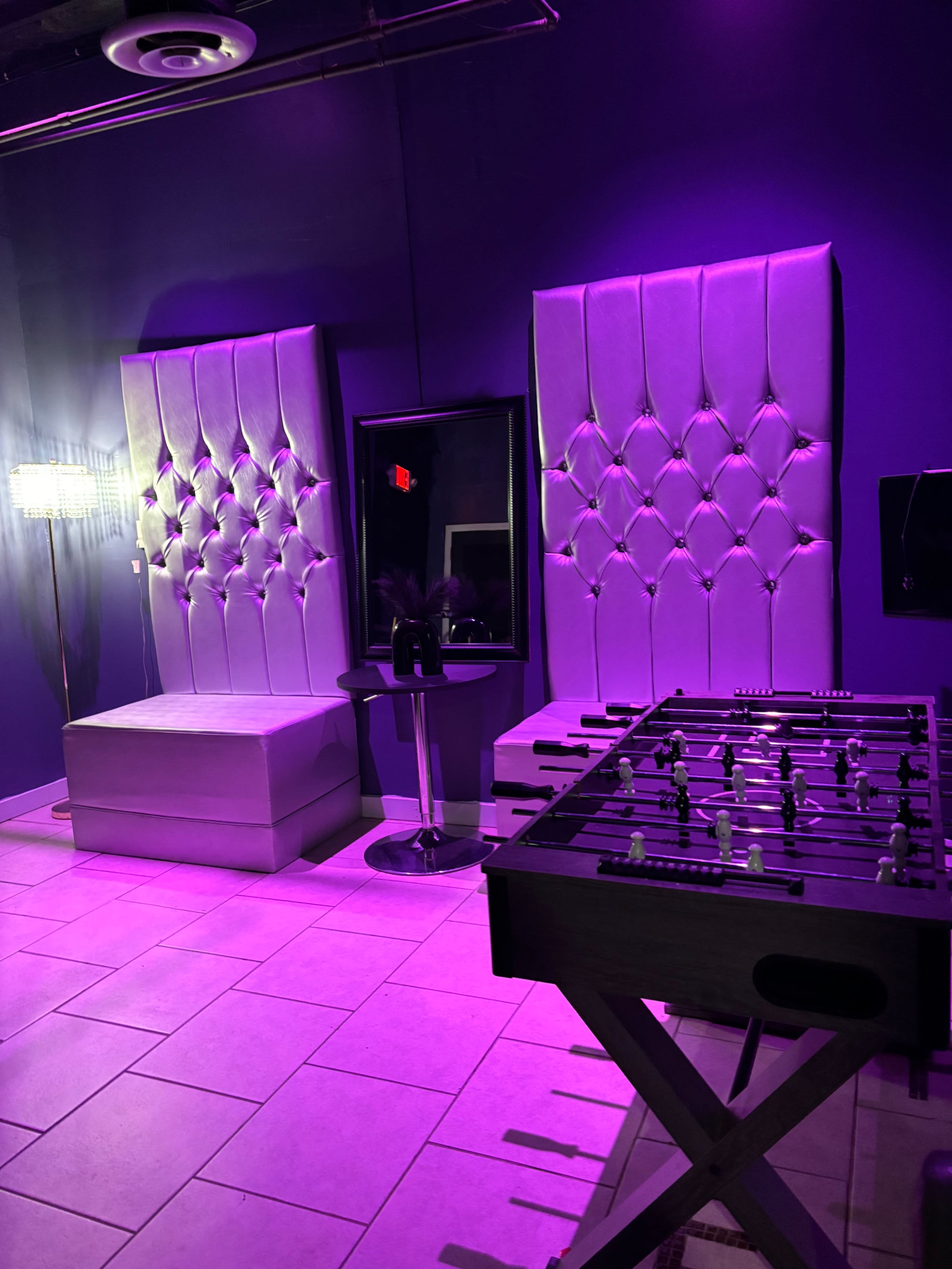 The image shows a modern lounge area featuring tufted seating, a small circular table, a mirror, and a foosball table, all illuminated by purple lighting.