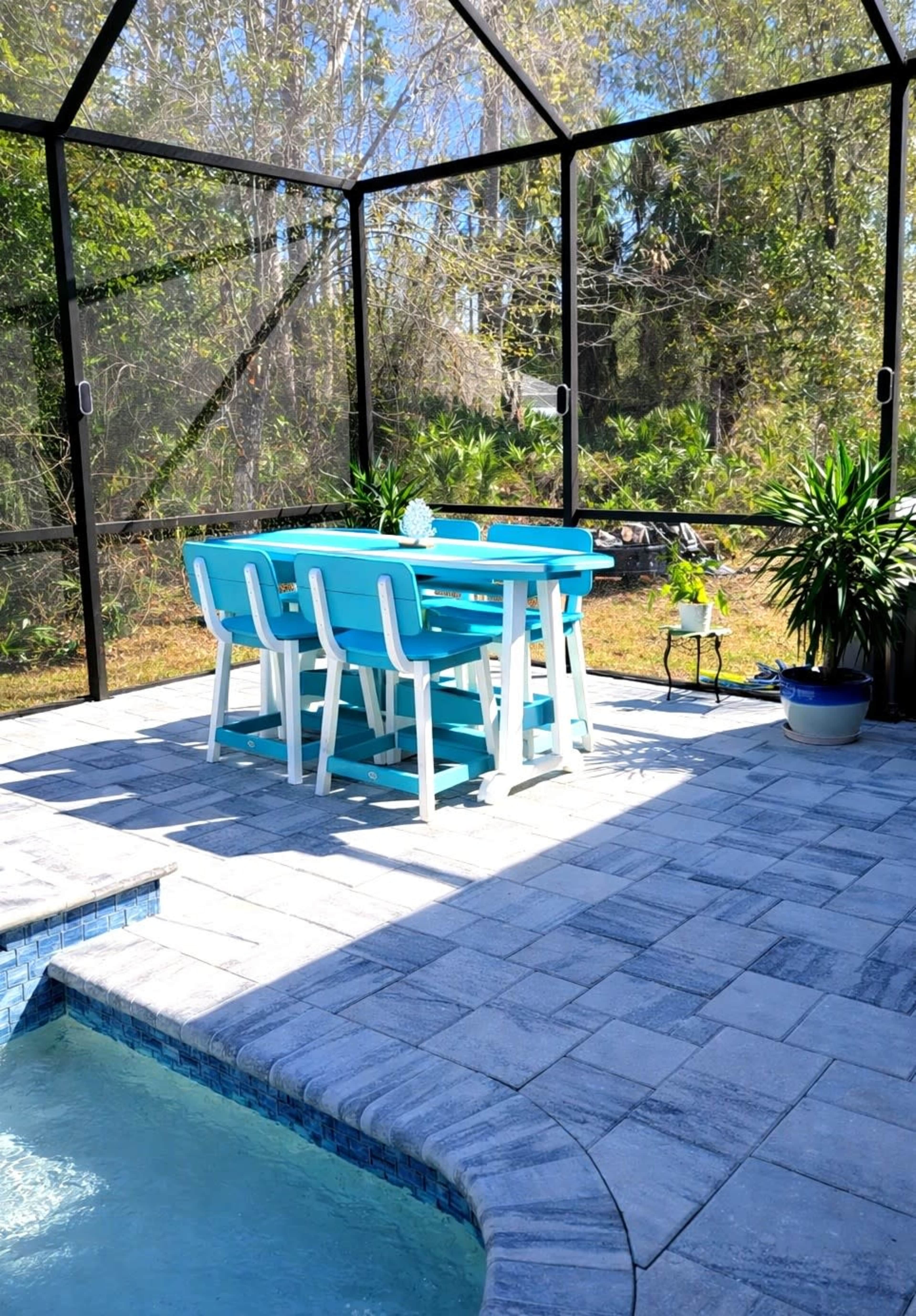 A turquoise dining table and chairs are set on a stone patio inside a screened enclosure, surrounded by greenery and featuring a pool to one side.