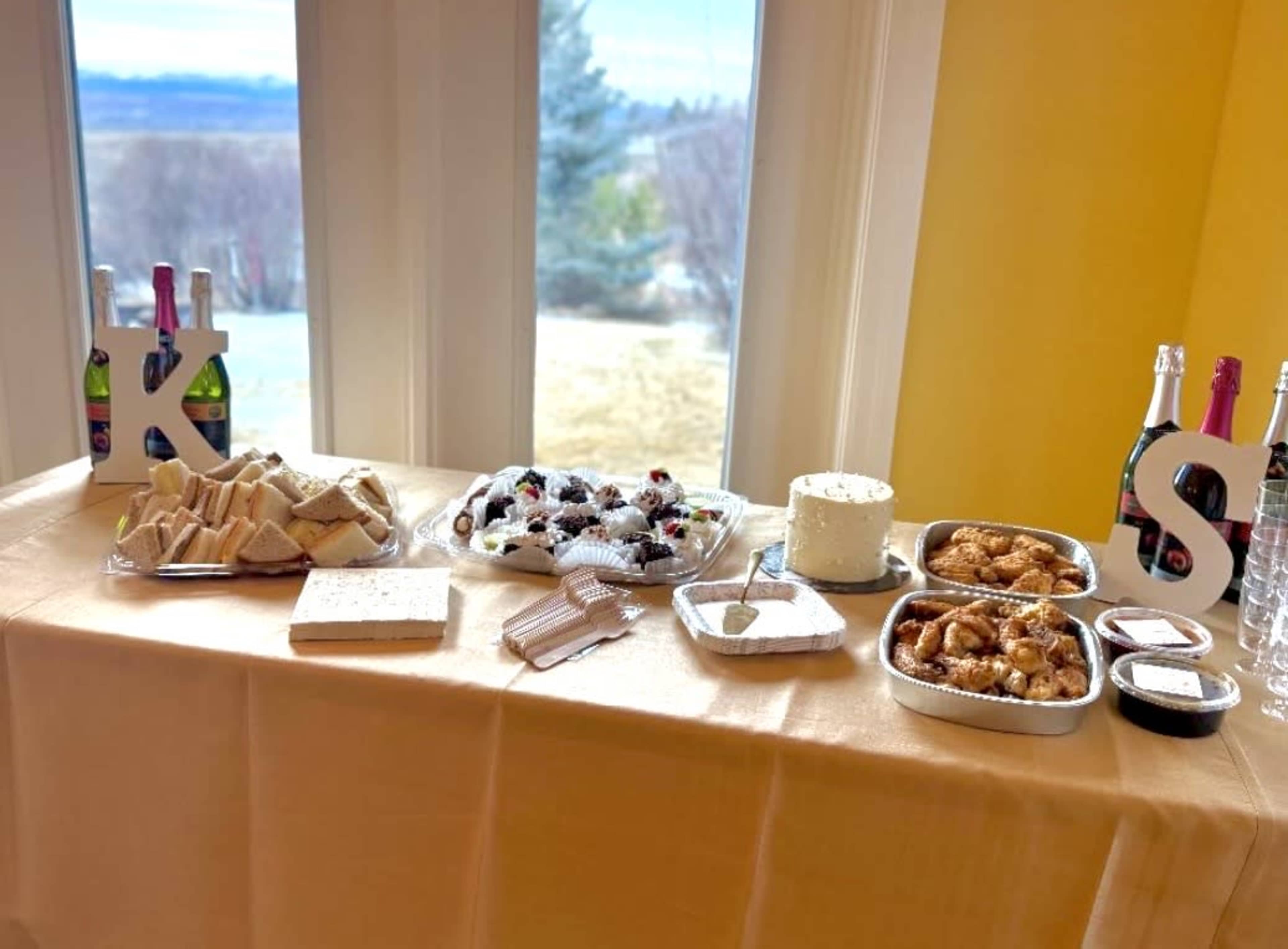 A table is set with various food items, including sandwiches, pastries, and a cake, alongside bottles of sparkling beverages, all arranged near large windows with a view.