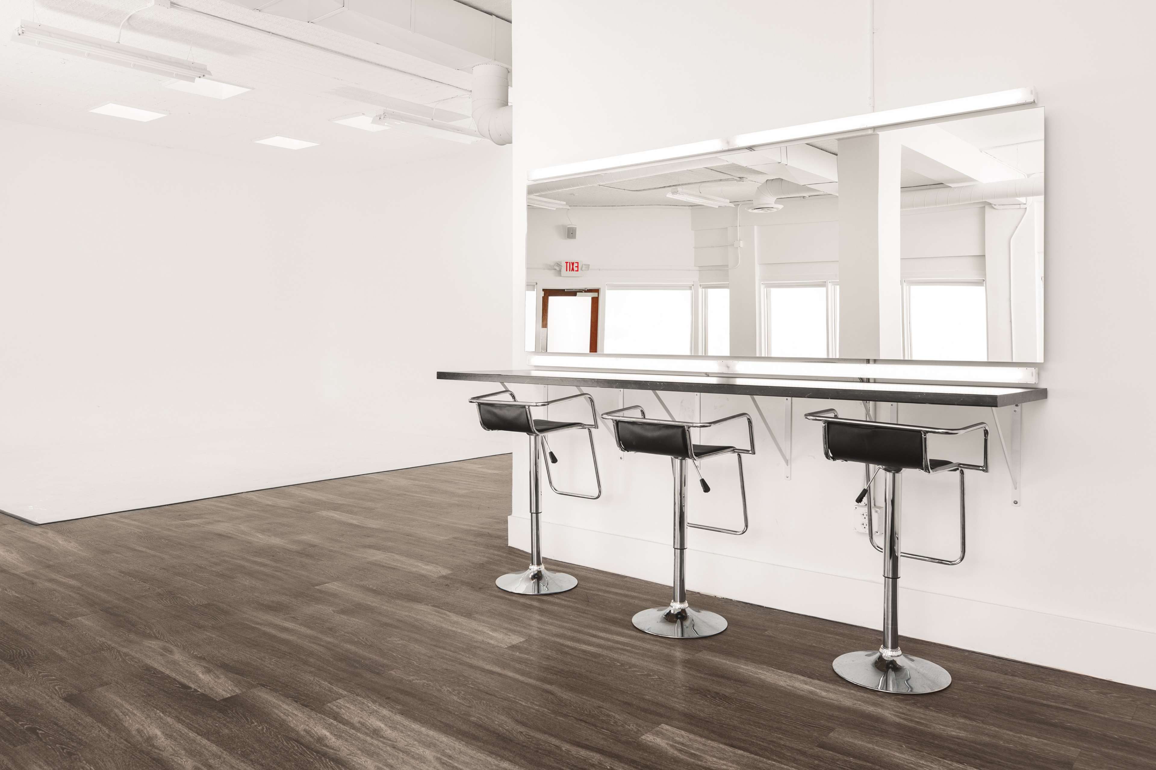 A minimalist interior with three black bar stools positioned at a counter facing a large mirror against a white wall.