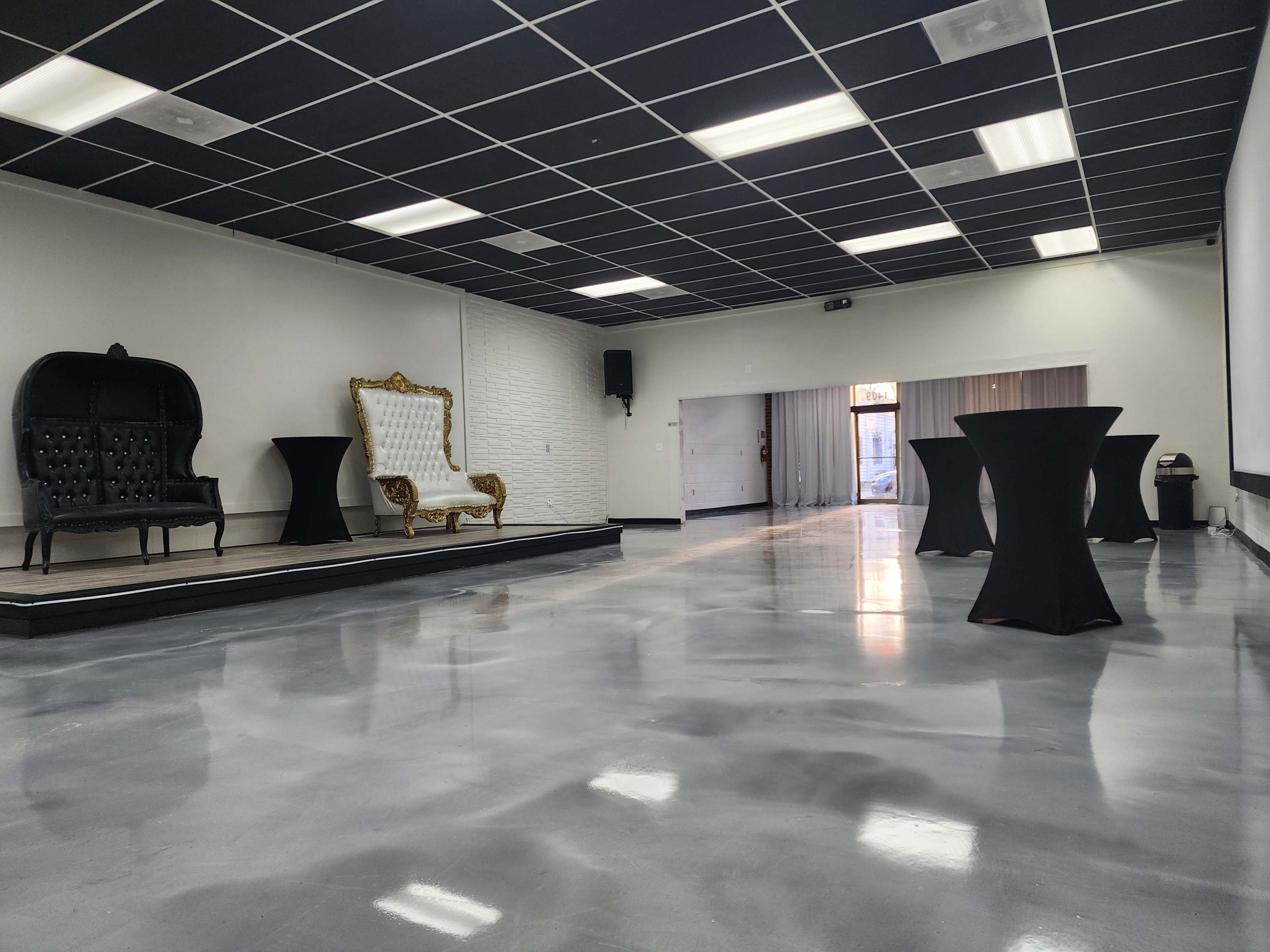 A spacious event room with polished concrete floors, two decorative chairs, and several black cocktail tables against a plain white wall.
