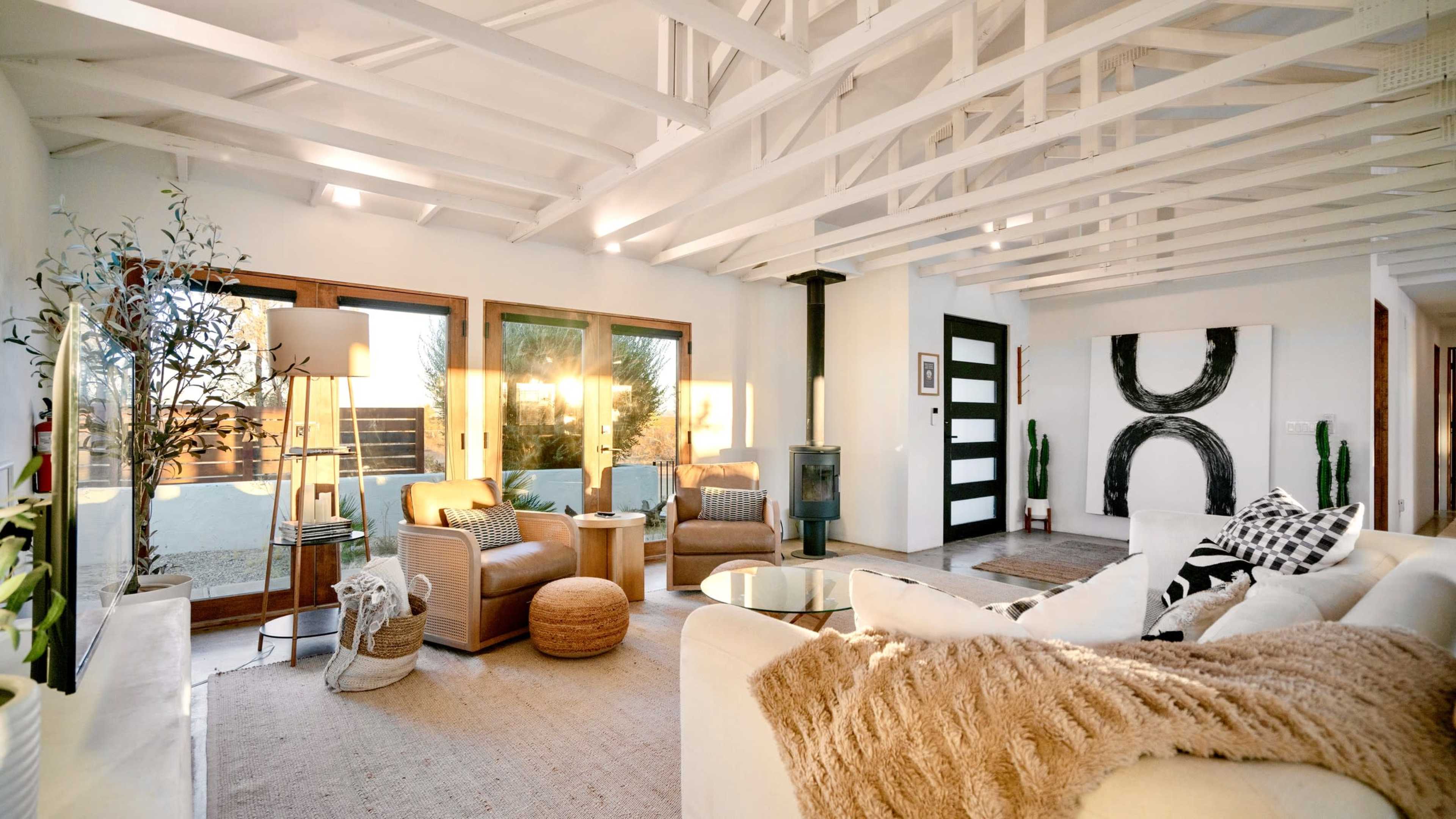 The image depicts a modern living room with large windows, two armchairs, a cozy sofa, and a minimalistic decor featuring a fireplace and plants.